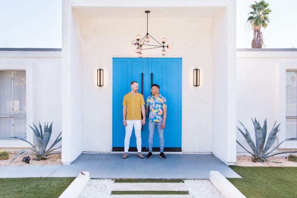 Two gay men standing in front of a mid-century modern architecture door that is bright blue in color. 