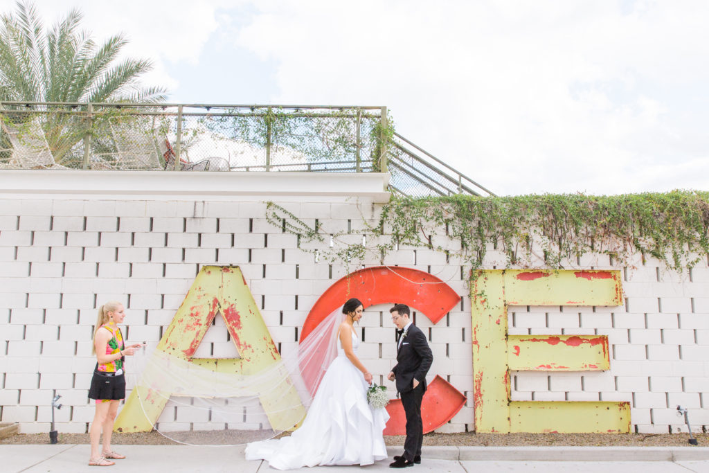 A photographer helping a bride with her vail. A bride and groom on their wedding day standing in front of steel letters spelling out ACE. 