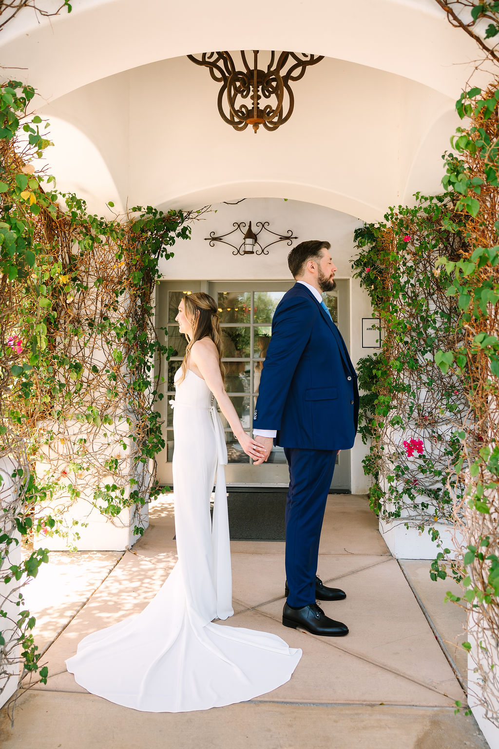 A bride and groom stand back to back holding hands for a first touch before their first look under an alcove at their alcazar palm springs wedding