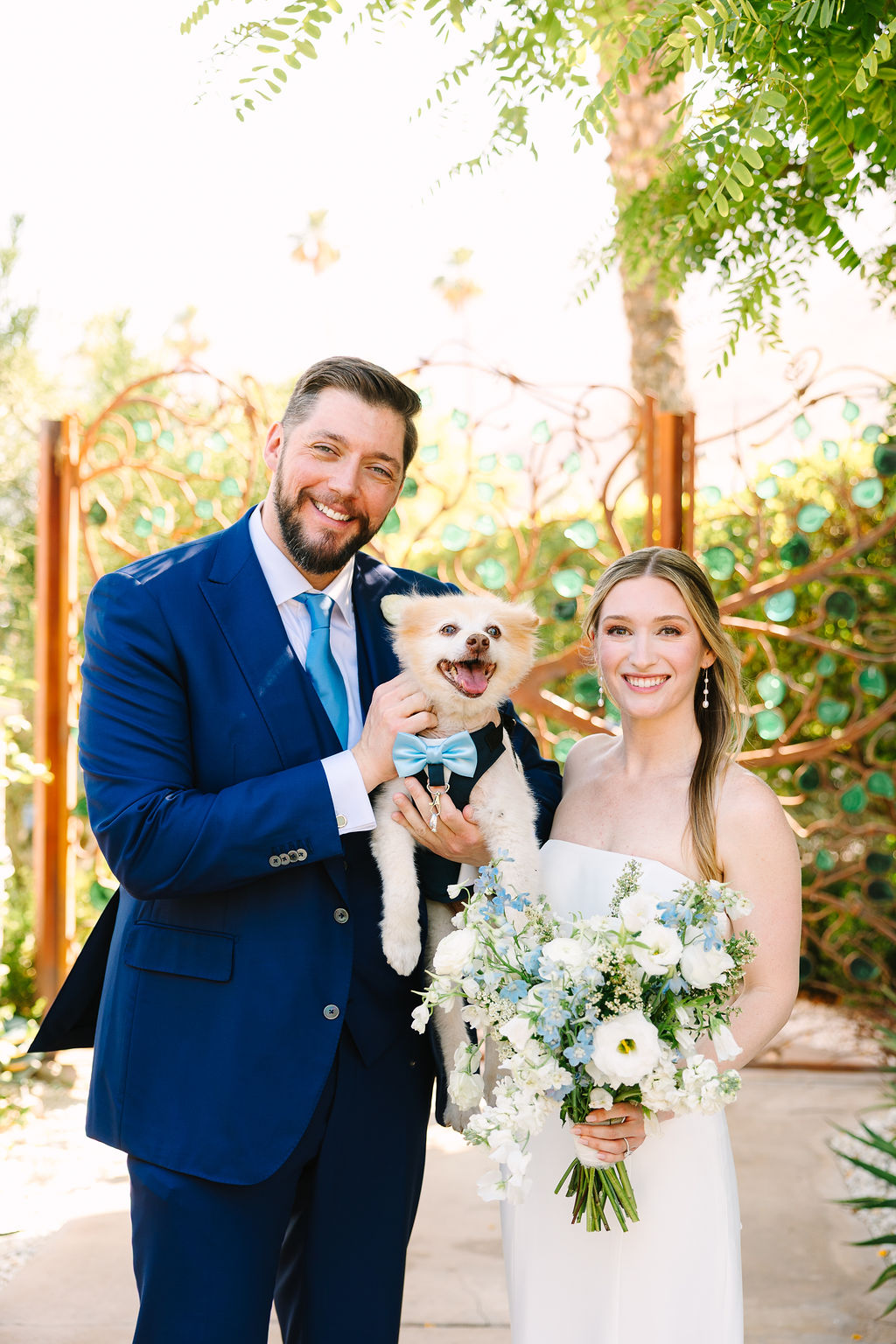 A bride and groom smile while standing with their tiny dog in a bowite