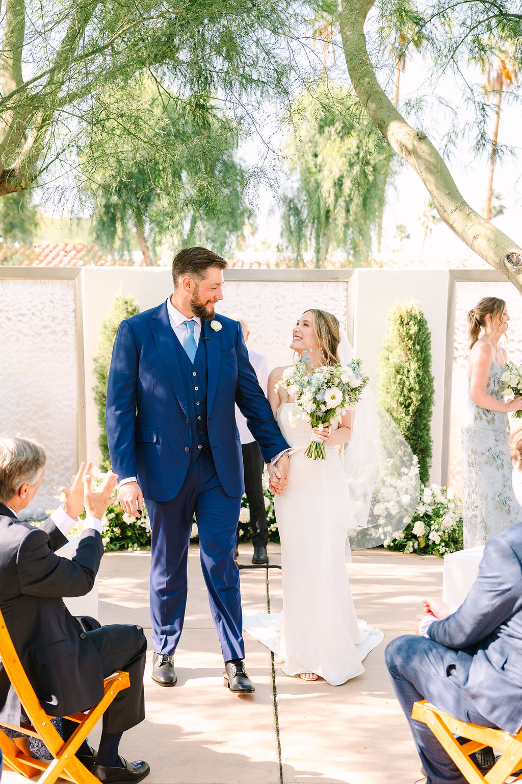 A bride and groom smile at each other while holding hands and walking up the aisle of the ceremony