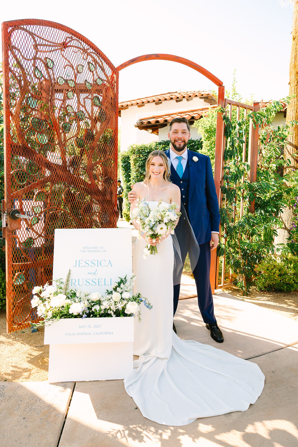 A bride and groom smile at the front gate to the alcazar palm springs wedding venue by their custom wedding sign