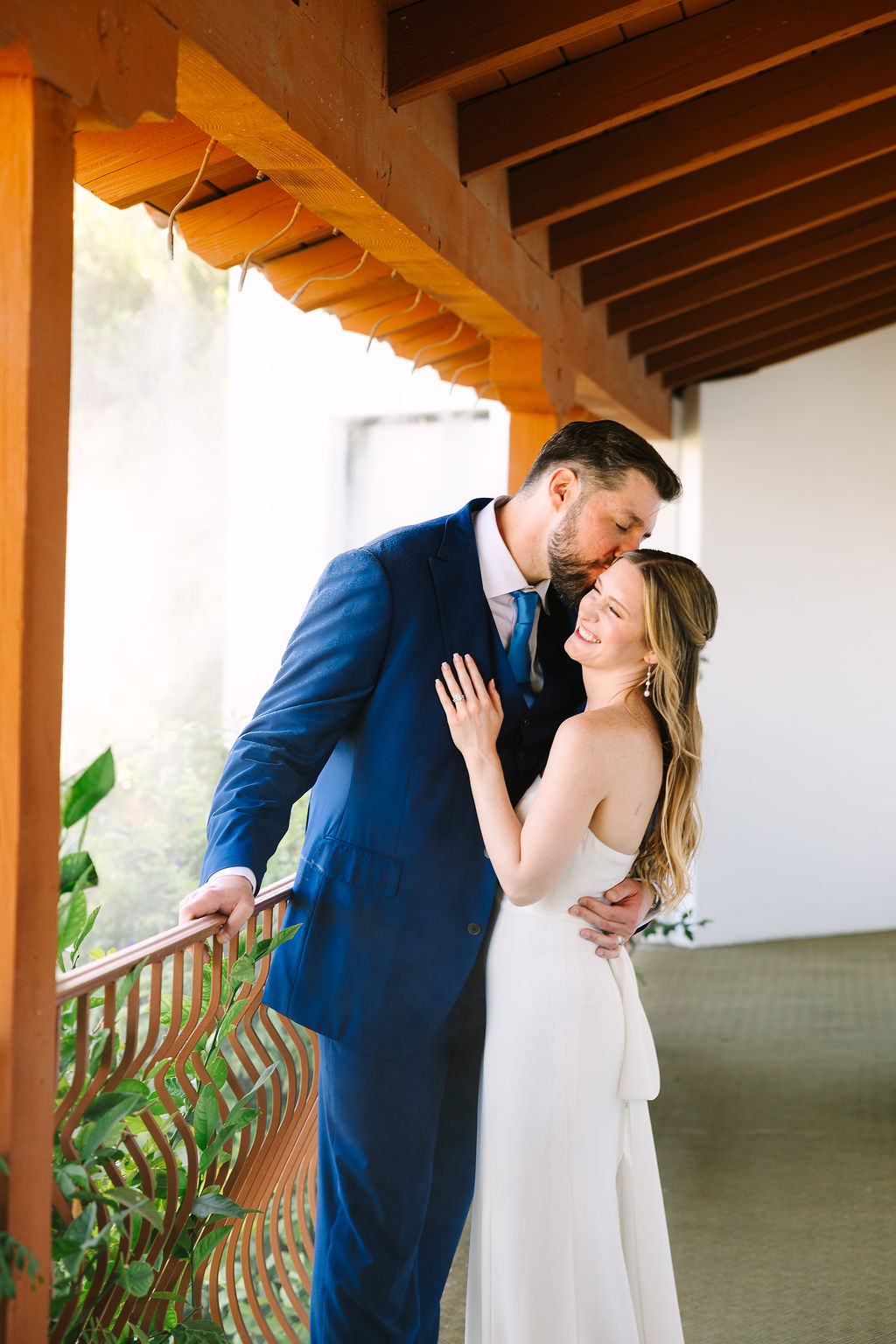 A bride smiles as her groom in a blue suit hugs and kisses her head on a porch