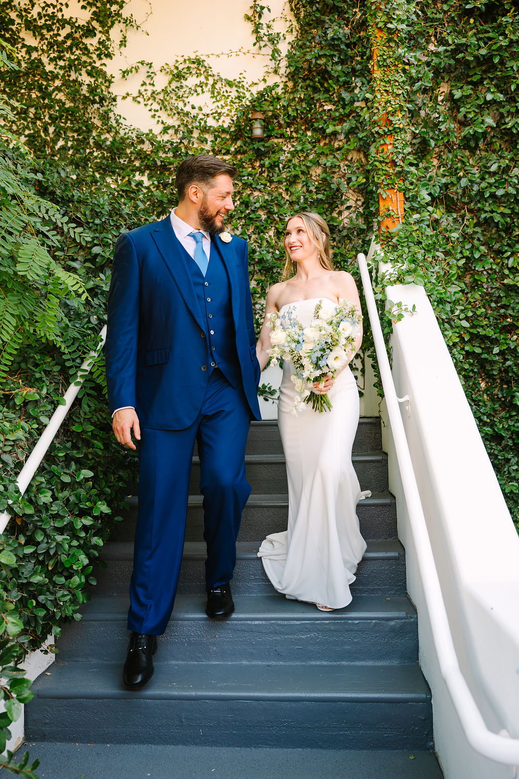 A groom walks down vine covered stairs holding hands and smiling at his bride at their alcazar palm springs wedding