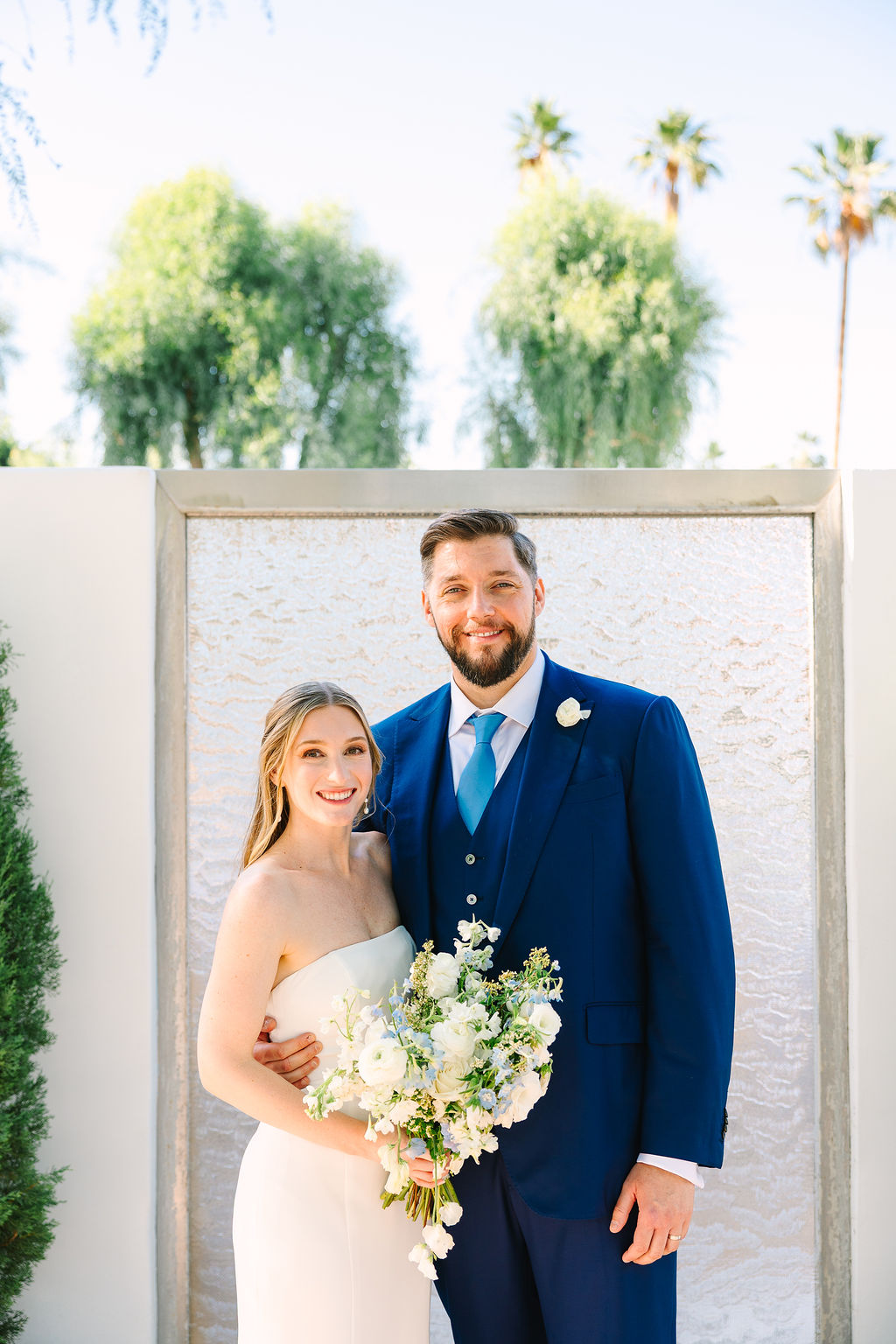 Newlyweds smile while hugging and holding the bouquet in front of a glass wall in a garden