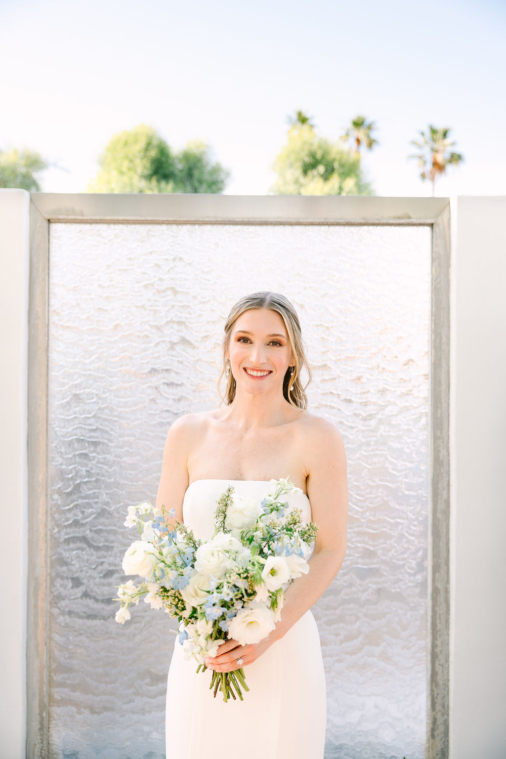 A bride smiles in front of a glass wall while holding her bouquet