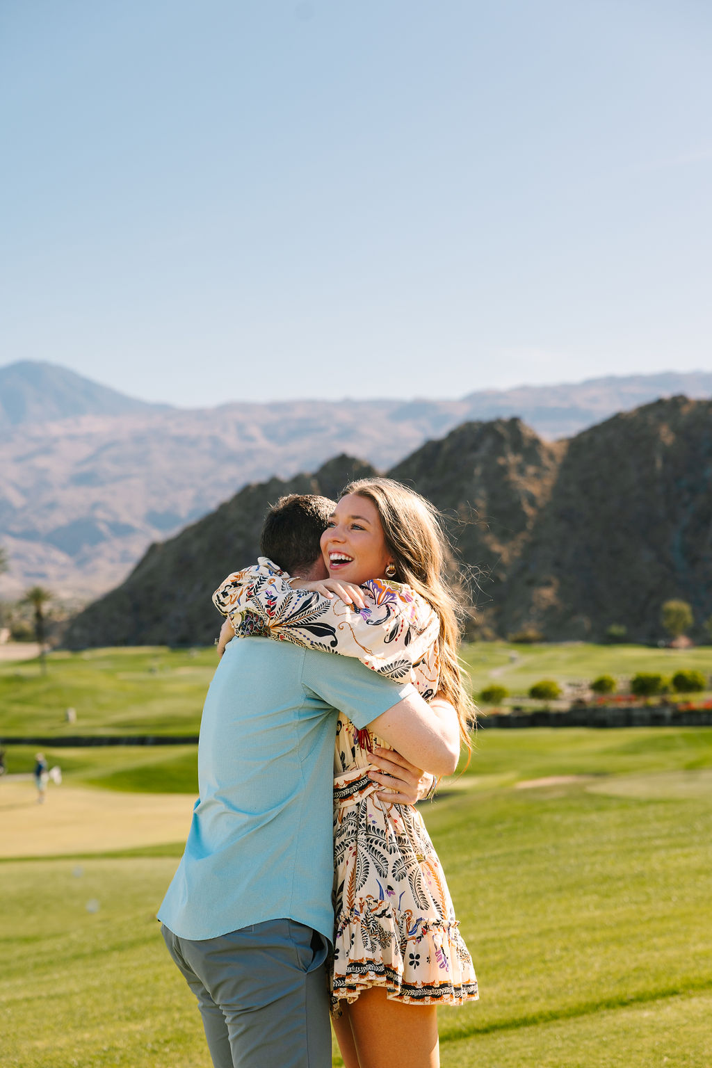 A woman embraces her fiancee right after saying yes to the ring on a golf course near the mountains