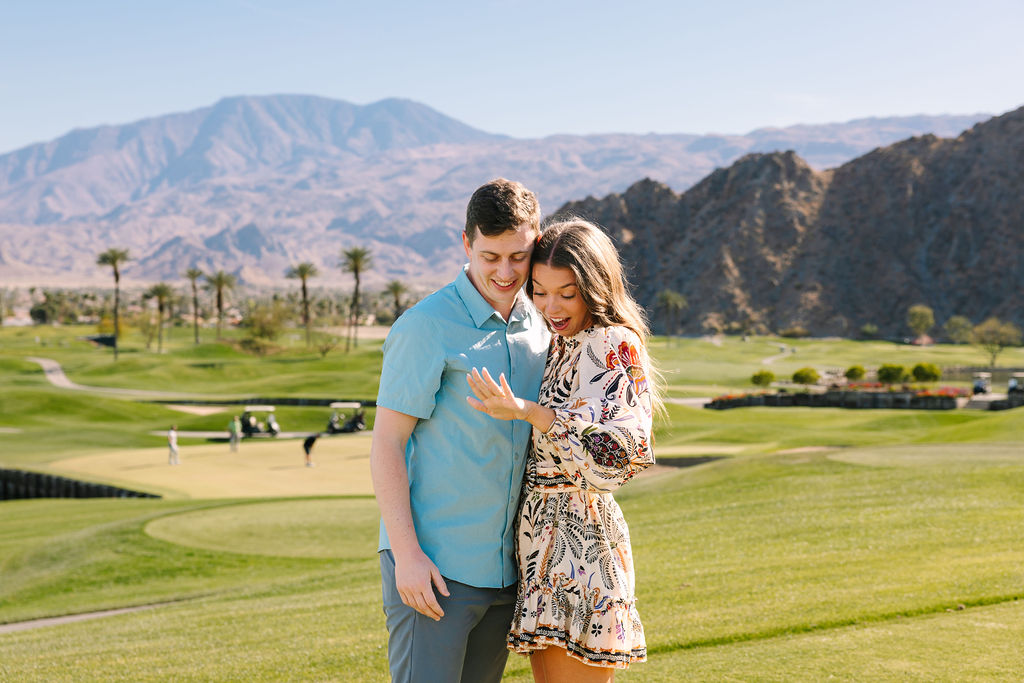 A happy newly engaged couple smile big while admiring the ring and touching heads on a golf green at pga west pete dye mountain course