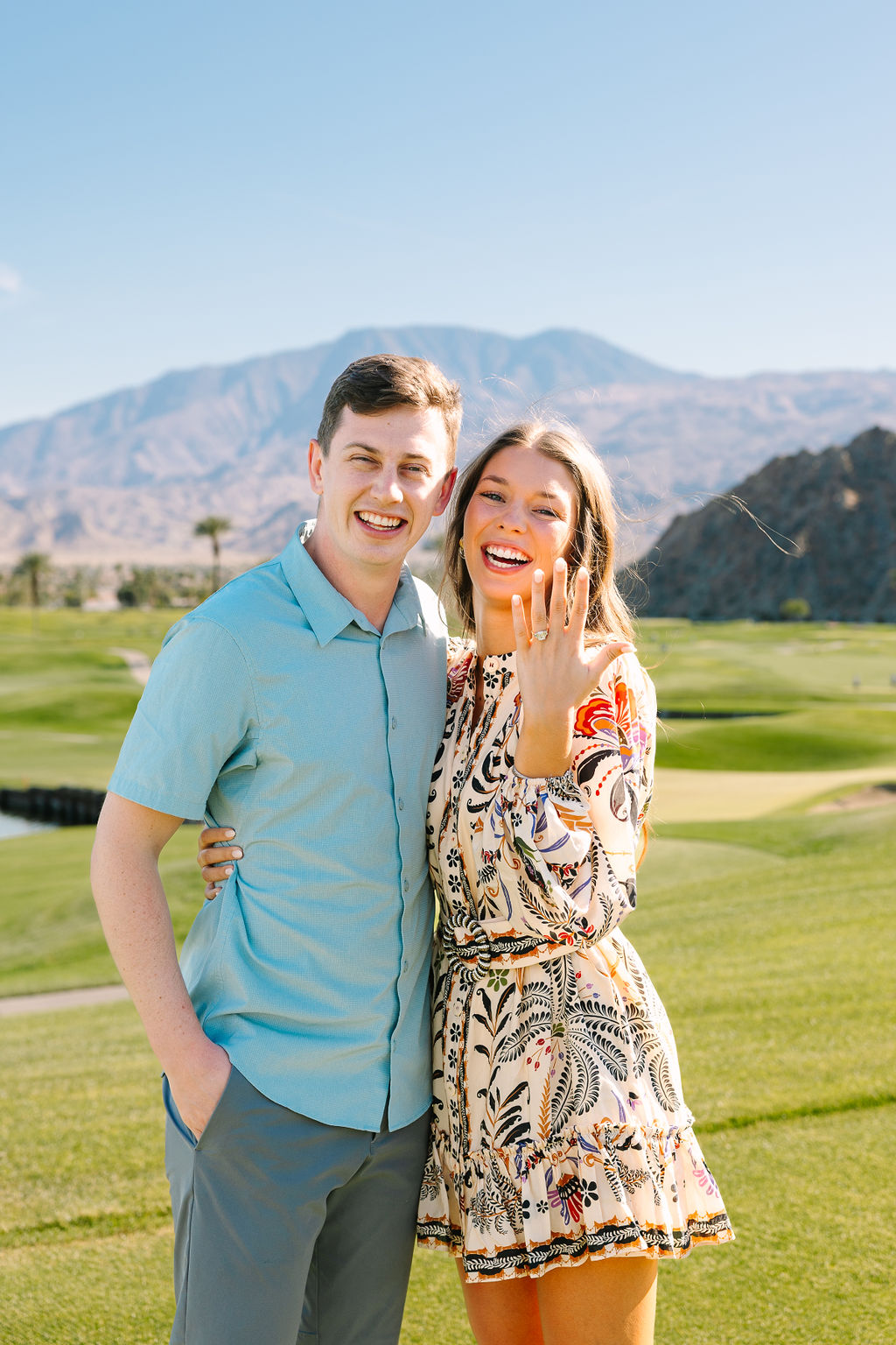 Happy newly engaged woman in a floral print dress shows off her ring with a big smile while hugging her fiancee in a blue shirt