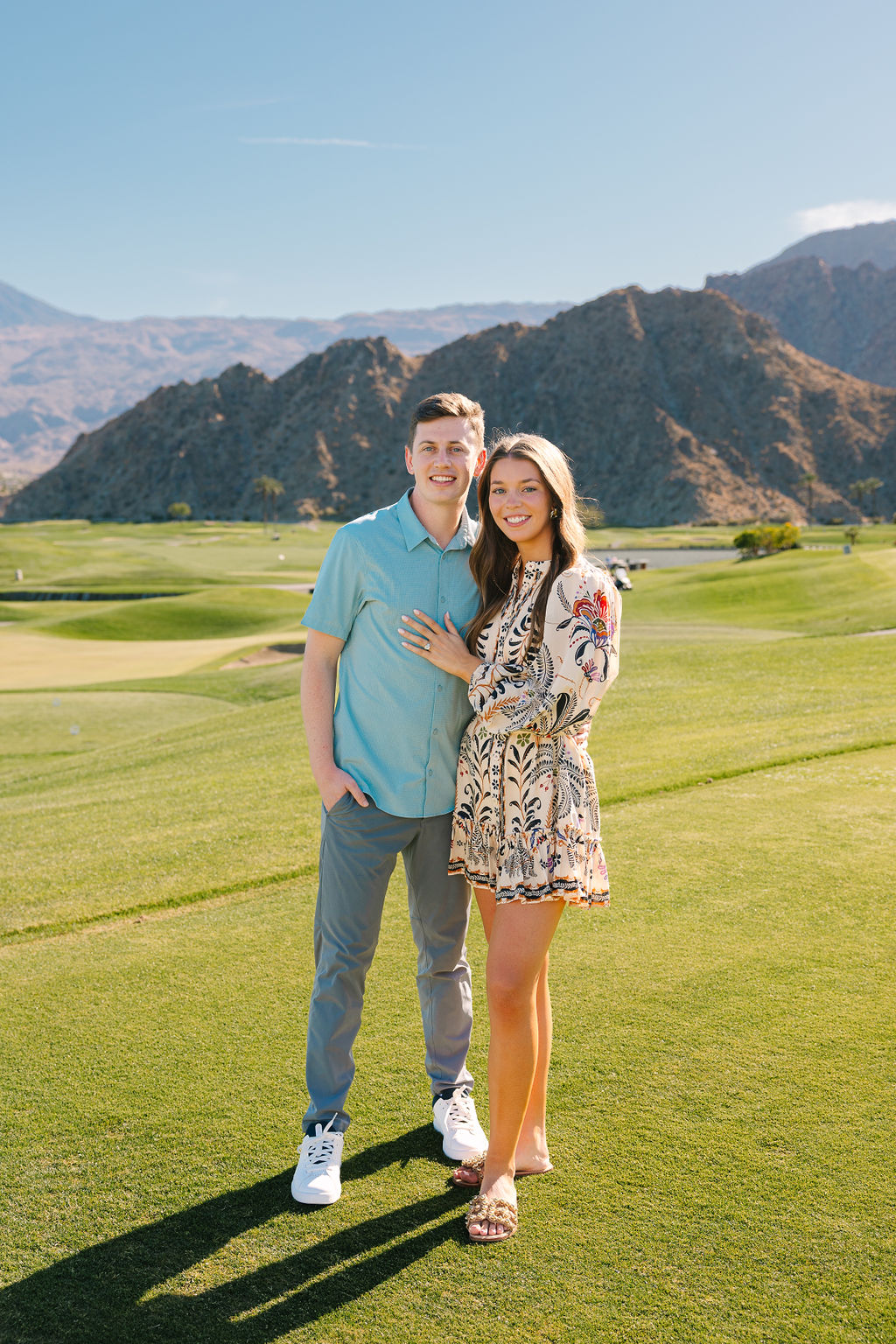 A happy engaged couple stand smiling on a pga west pete dye mountain course putting green with mountains in the background