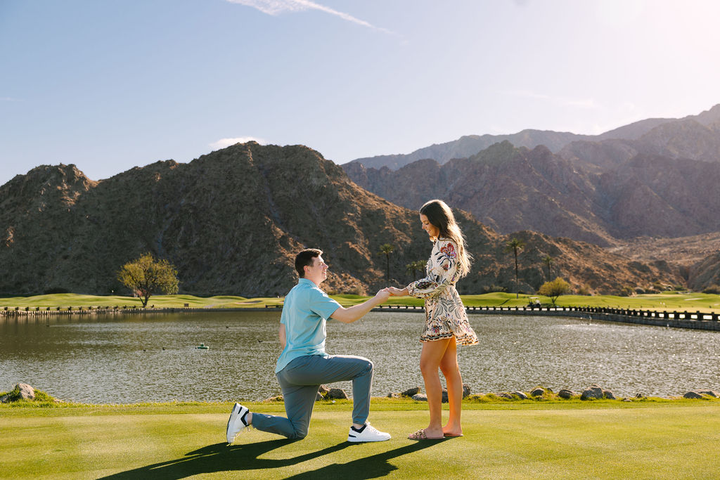 A man kneels on a putting green by a pond after proposing to his girlfriend in a colorful dress at pga west pete dye mountain course