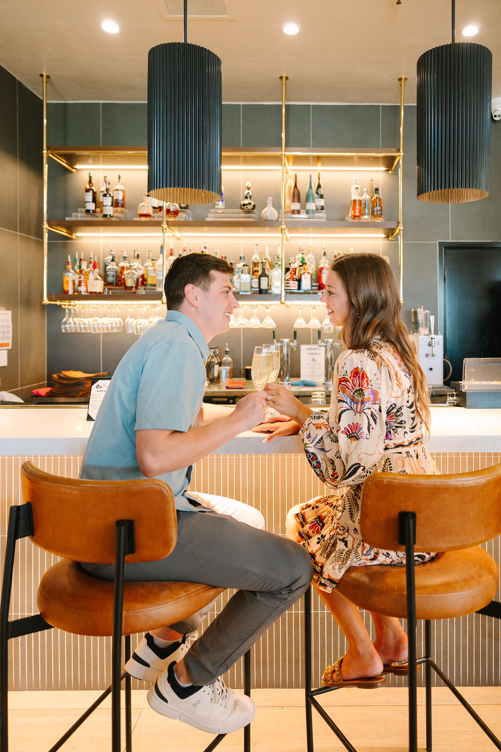 A happy engaged couple toast champagne while sitting at a bar at pga west pete dye mountain course in a blue polo and colorful dress