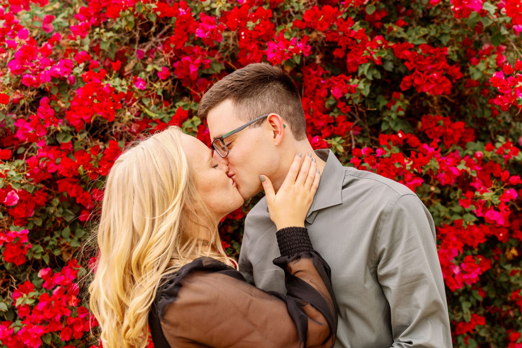 A happy newly engaged couple in a black dress and grey button down share a kiss in front of pink wall of flowers at the kimpton rowan palm springs hotel