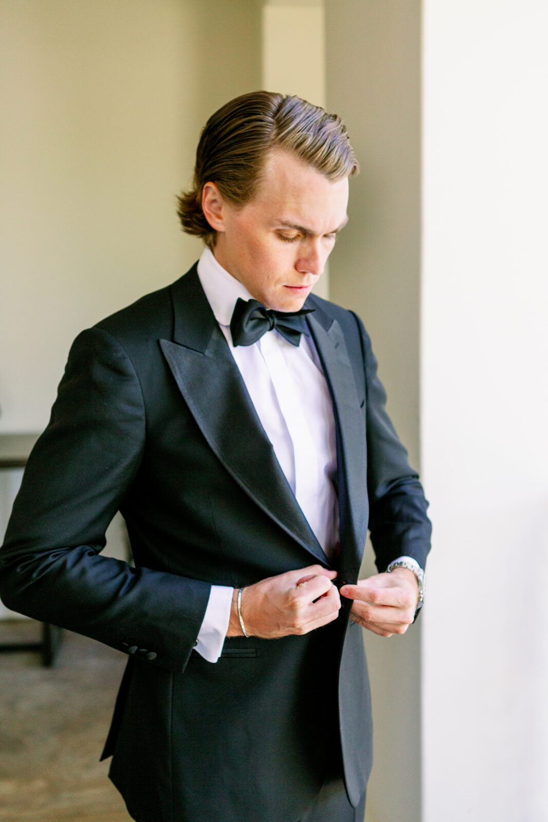 A groom buttons his tuxedo jacket while standing in a window