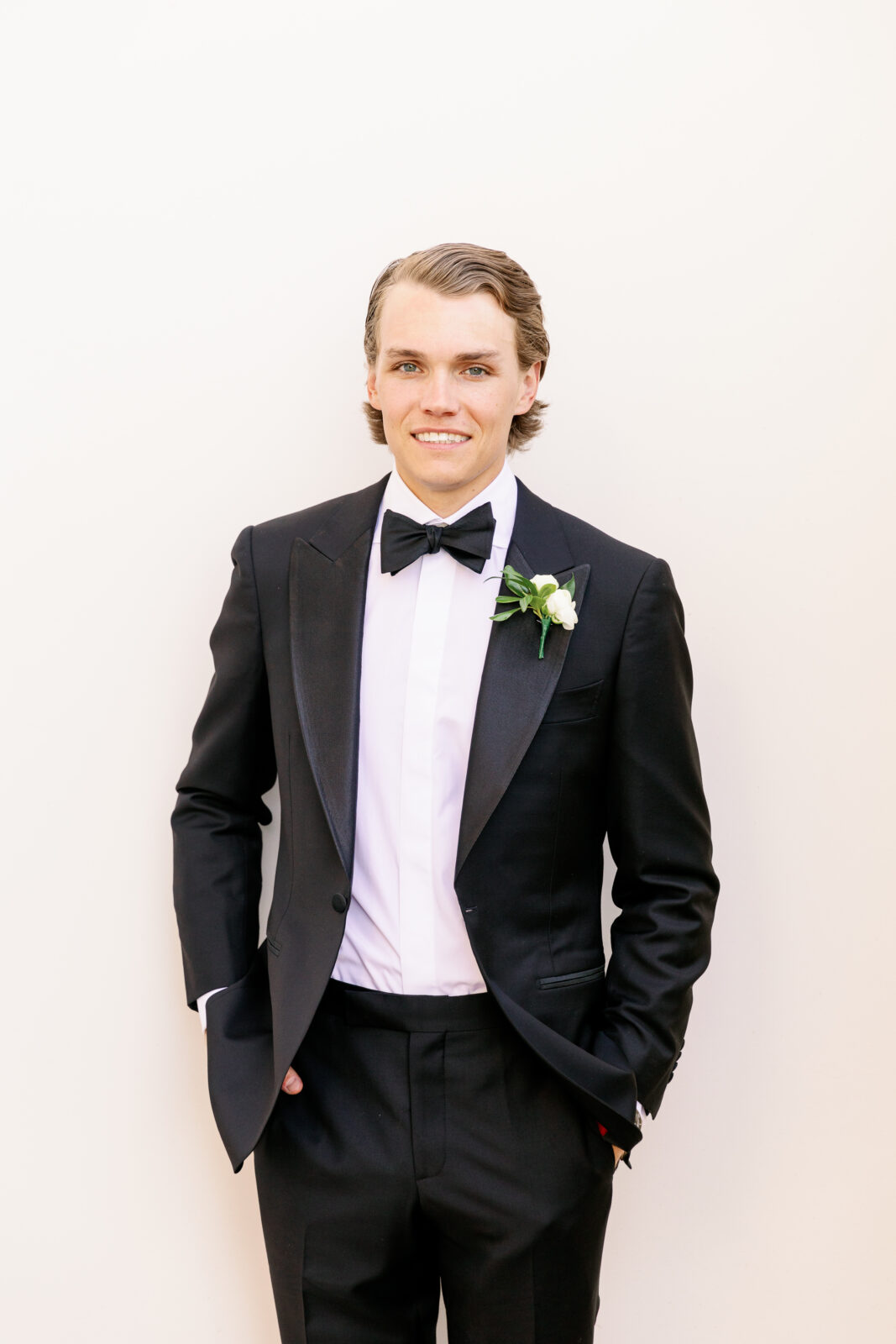 A groom in a black tuxedo stands against a wall with hands in pockets