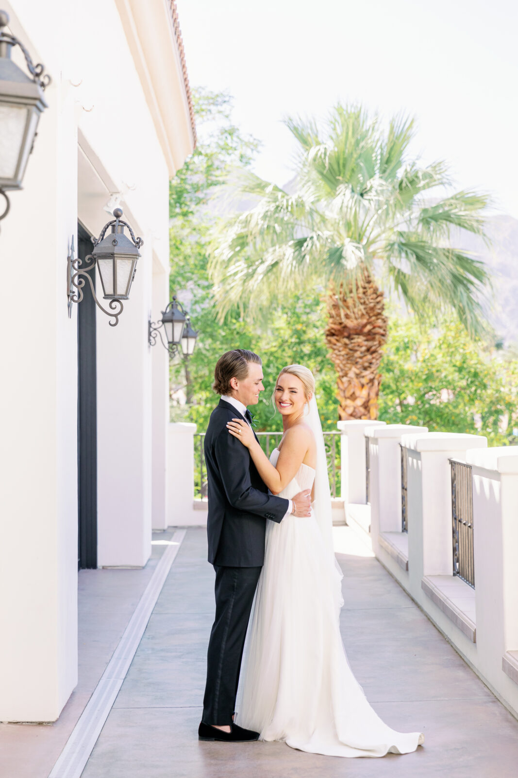 A bride smiles over her shoulder while hugging her groom on a balcony at la quinta country club wedding venue