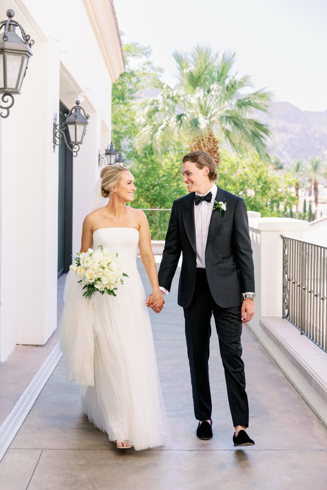 A bride and groom smile while walking hand in hand on the balcony