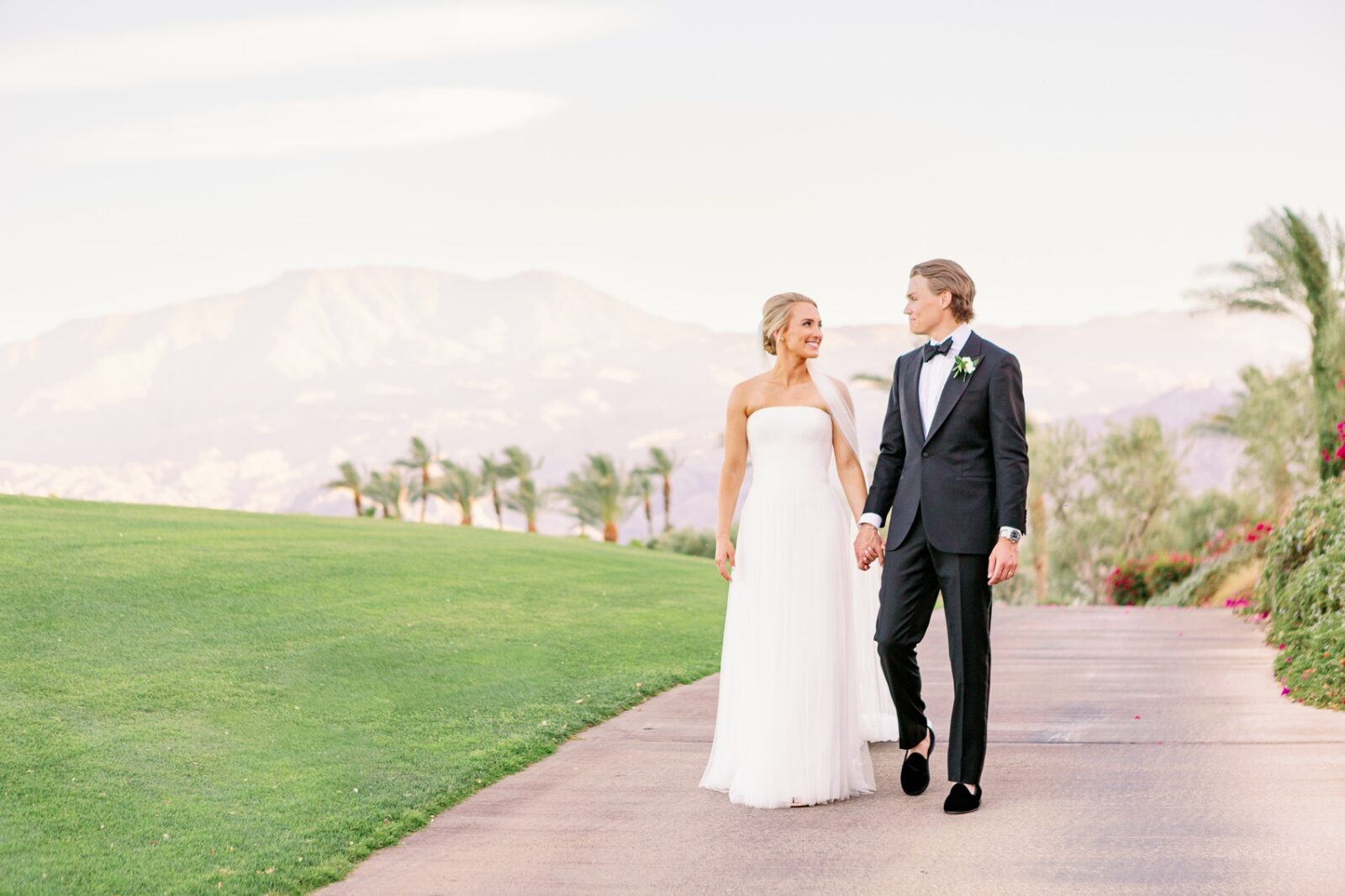 A bride and groom smile at each other while walking holding hands on a sidewalk at their la quinta country club wedding