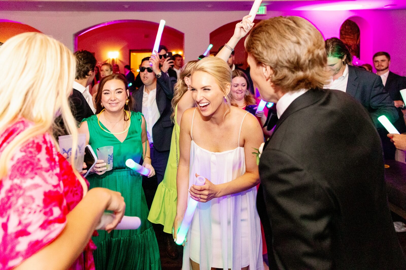 A bride and groom laugh and dance during their reception with guests and glow sticks