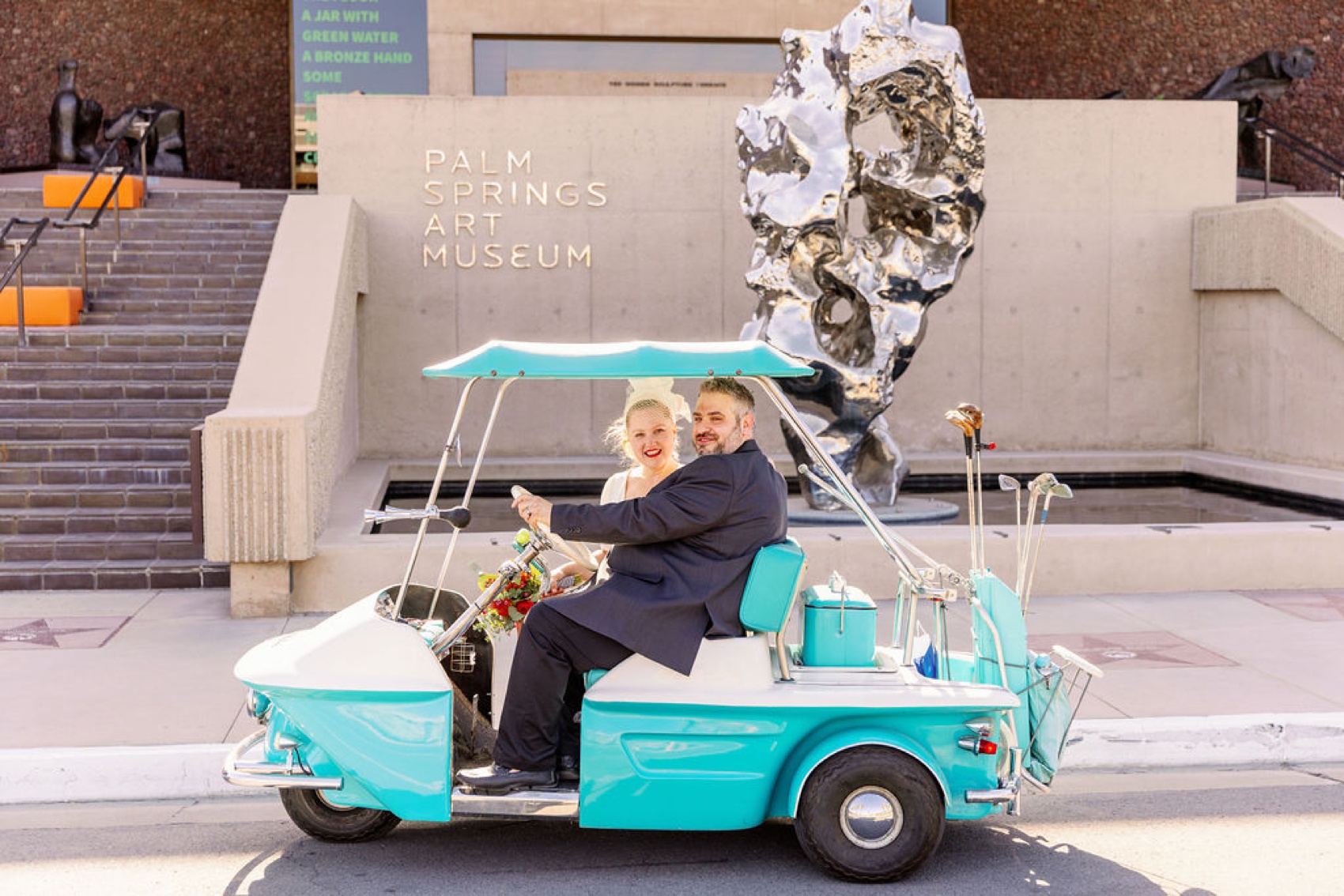 A bride and groom sit in a vintage blue golf cart in front of the palm springs art museum wedding venue