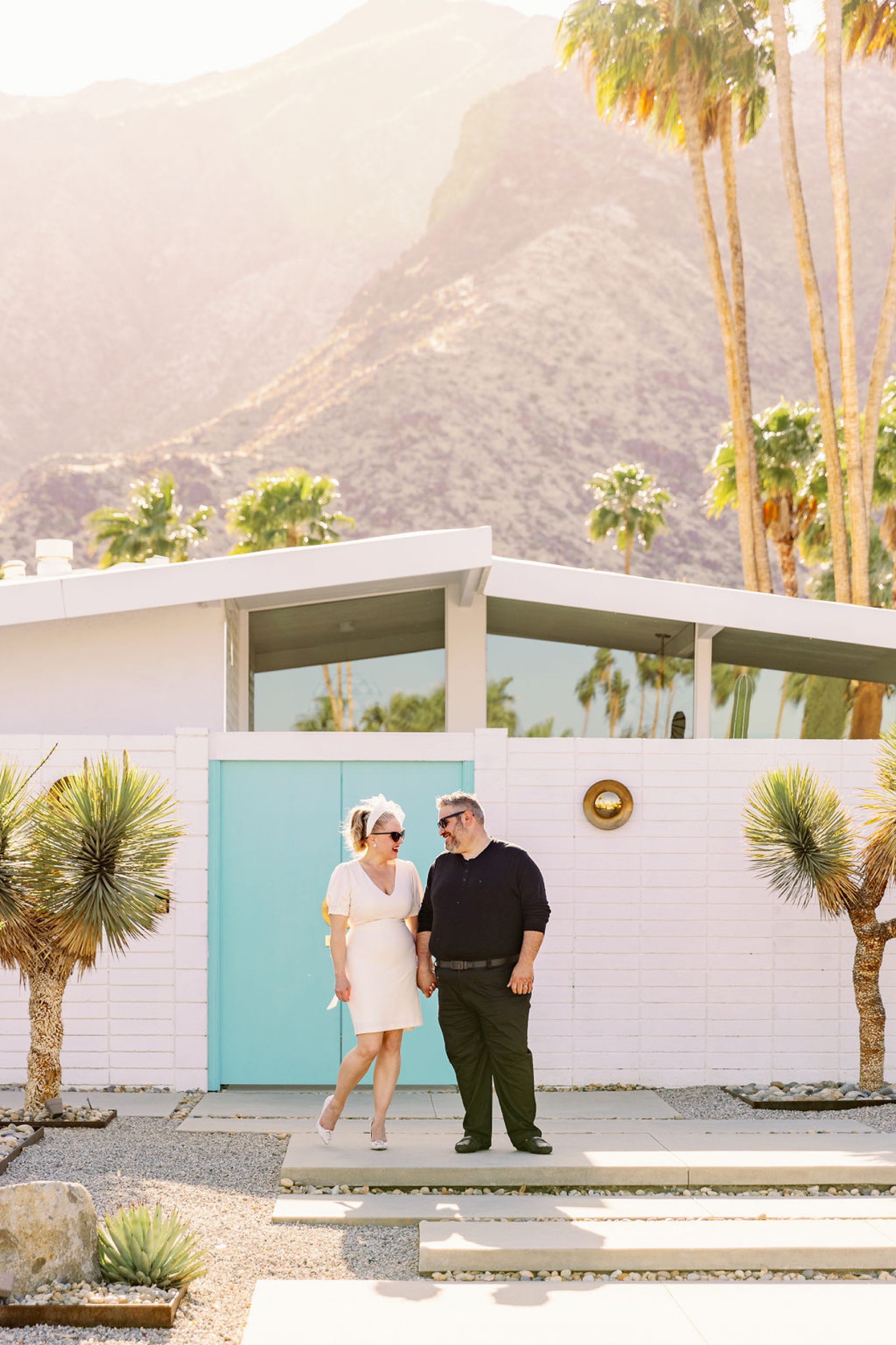 A happy couple hold hands while laughing together in front of a mid century modern building in the desert at sunset