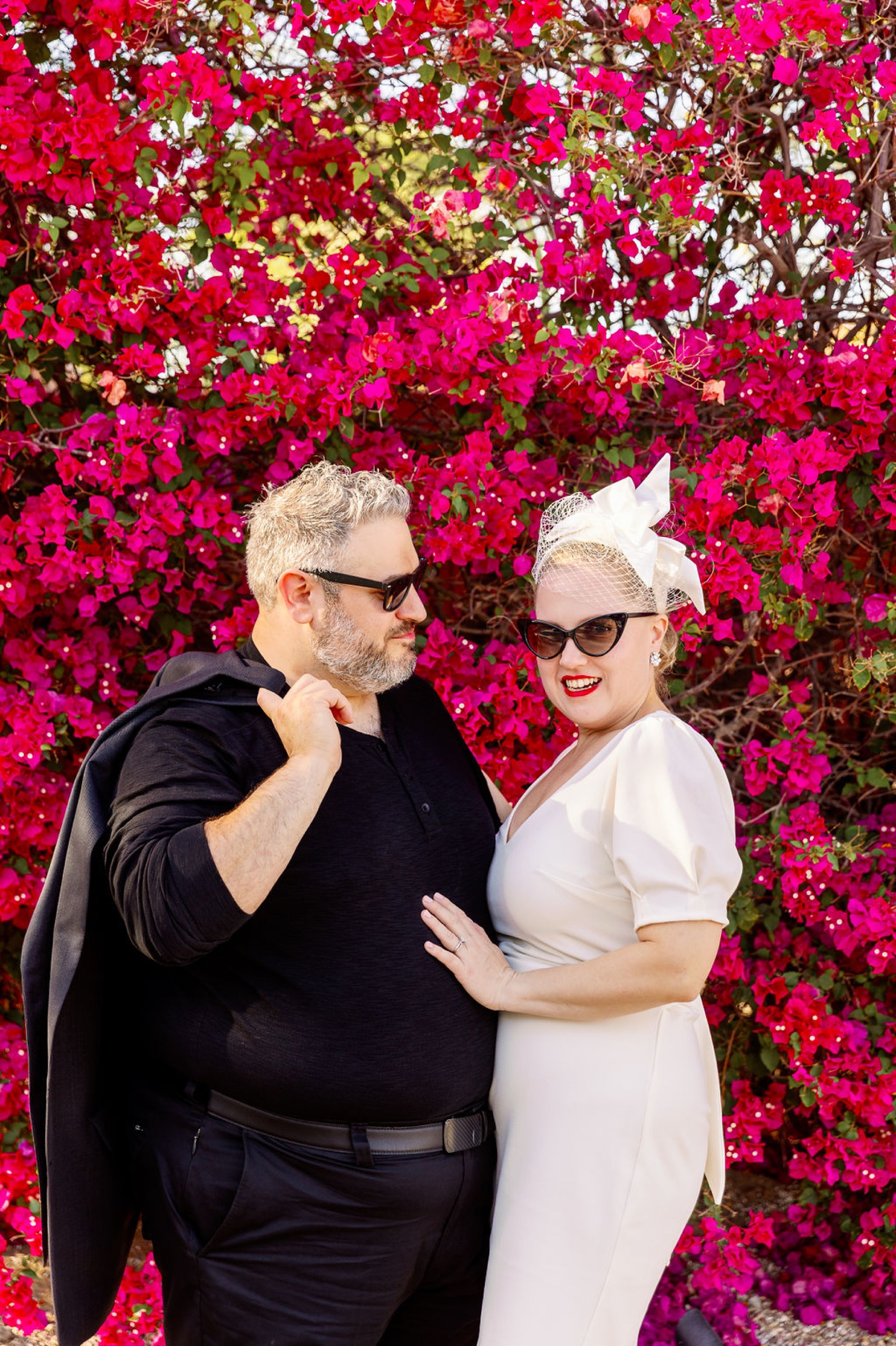 A bride and groom stand smiling in front of vibrant pink flowers at their palm springs art museum wedding