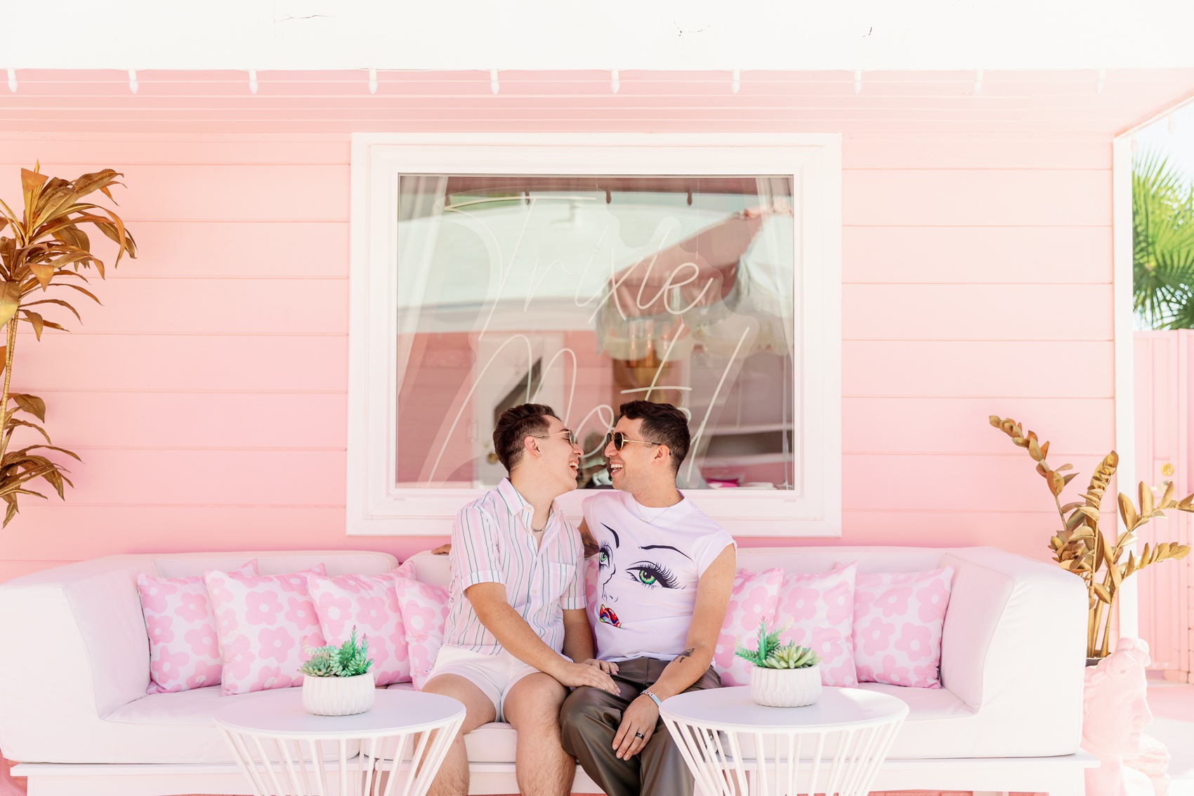 A happy engaged couple in pink shirts sit on a matching couch under the neon motel sign while laughing at each other