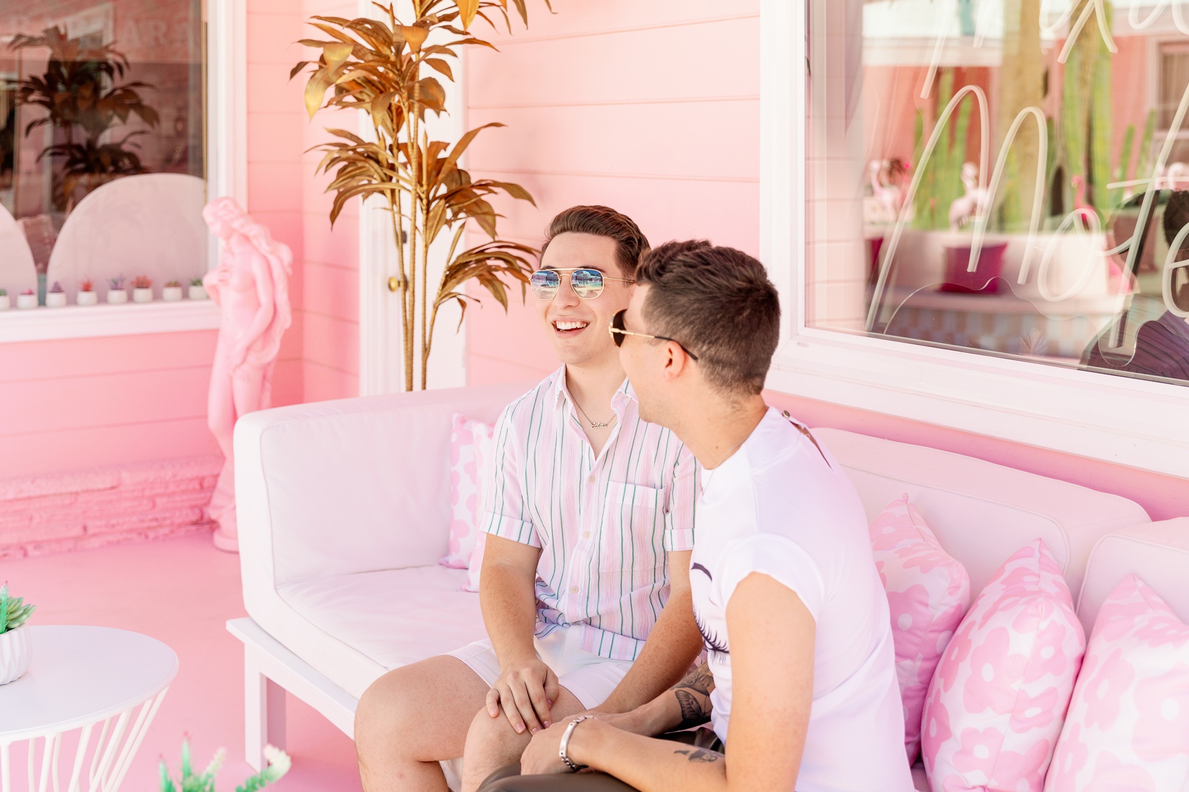 A couple share a laugh after getting engaged on a pink patio couch in matching shirts and sunglasses