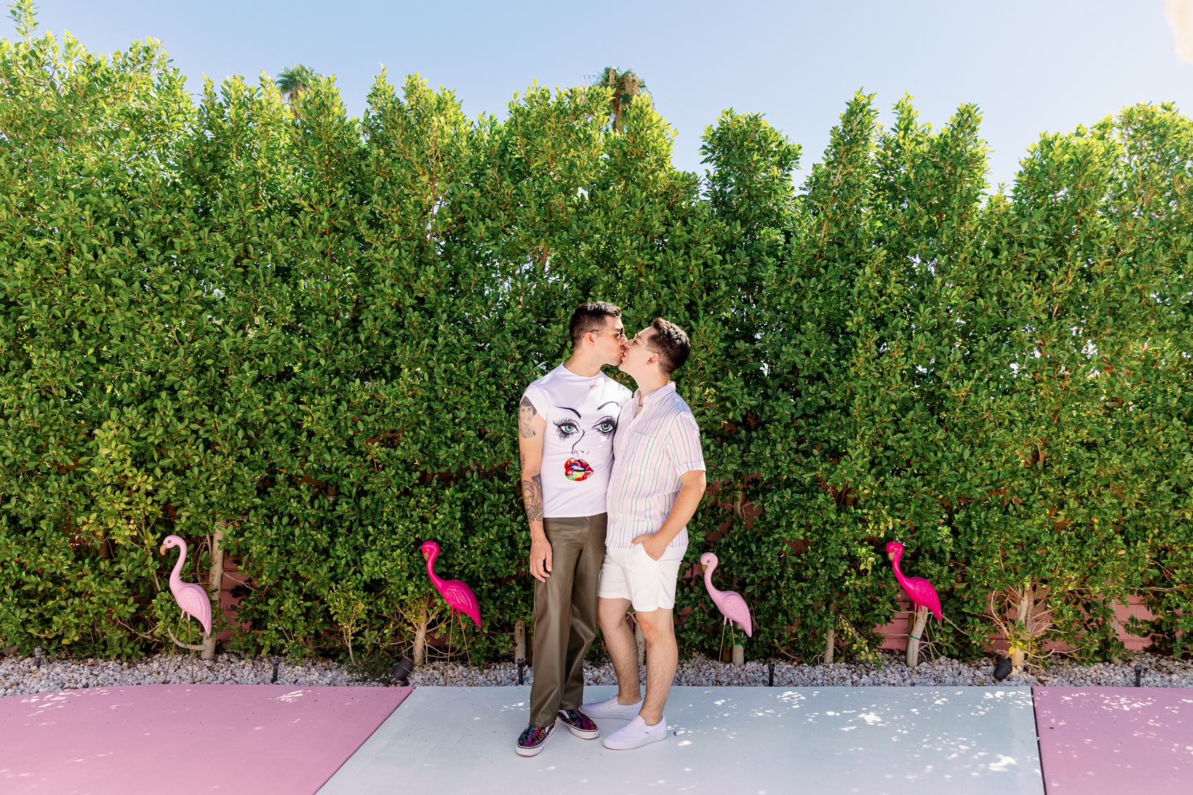 A couple kisses after getting engaged and staying at the trixie motel palm springs against a hedge wall with pink flamingos