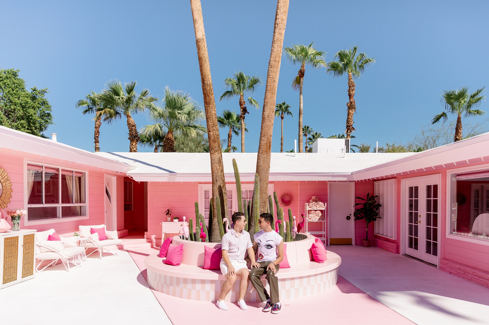 An engaged couple chat and laugh while sitting on a planter bench in pink shirts to match the trixie motel palm springs
