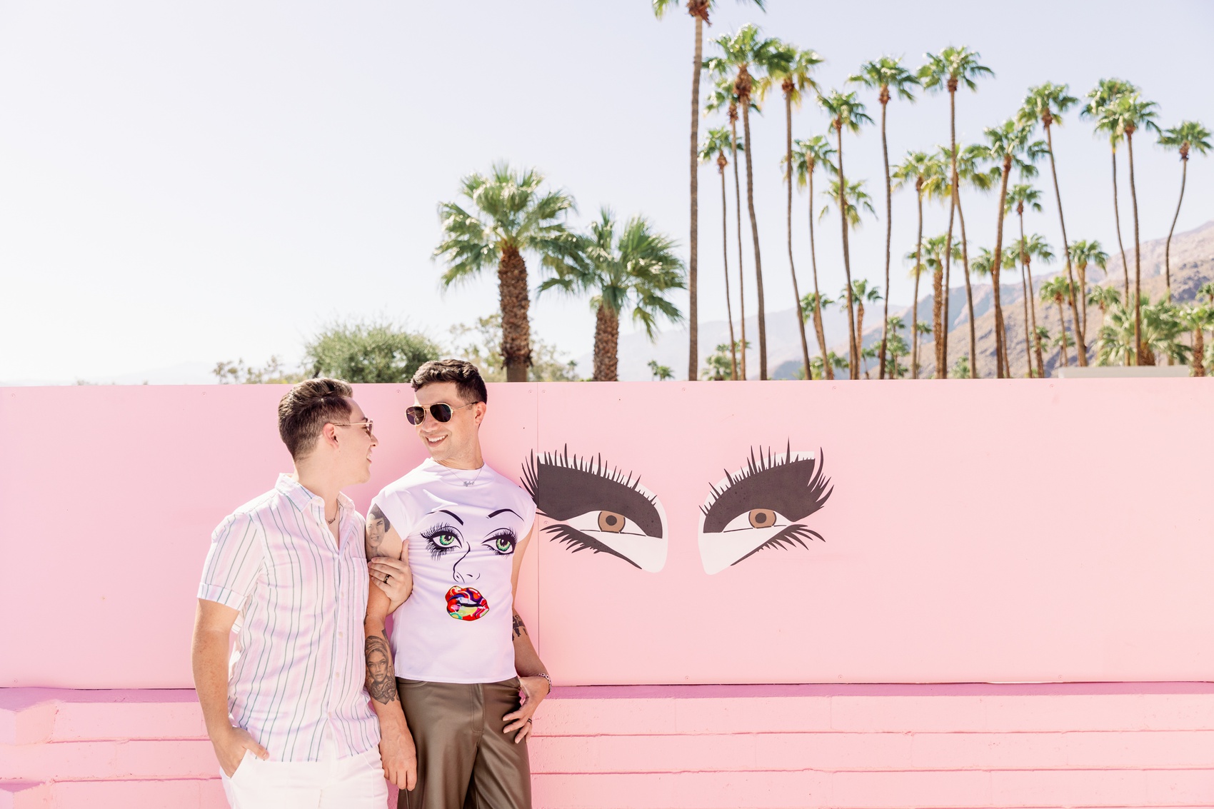 An engaged couple in pink shirts smile at each other while leaning on a pink wall with an eyes mural at the trixie motel palm springs