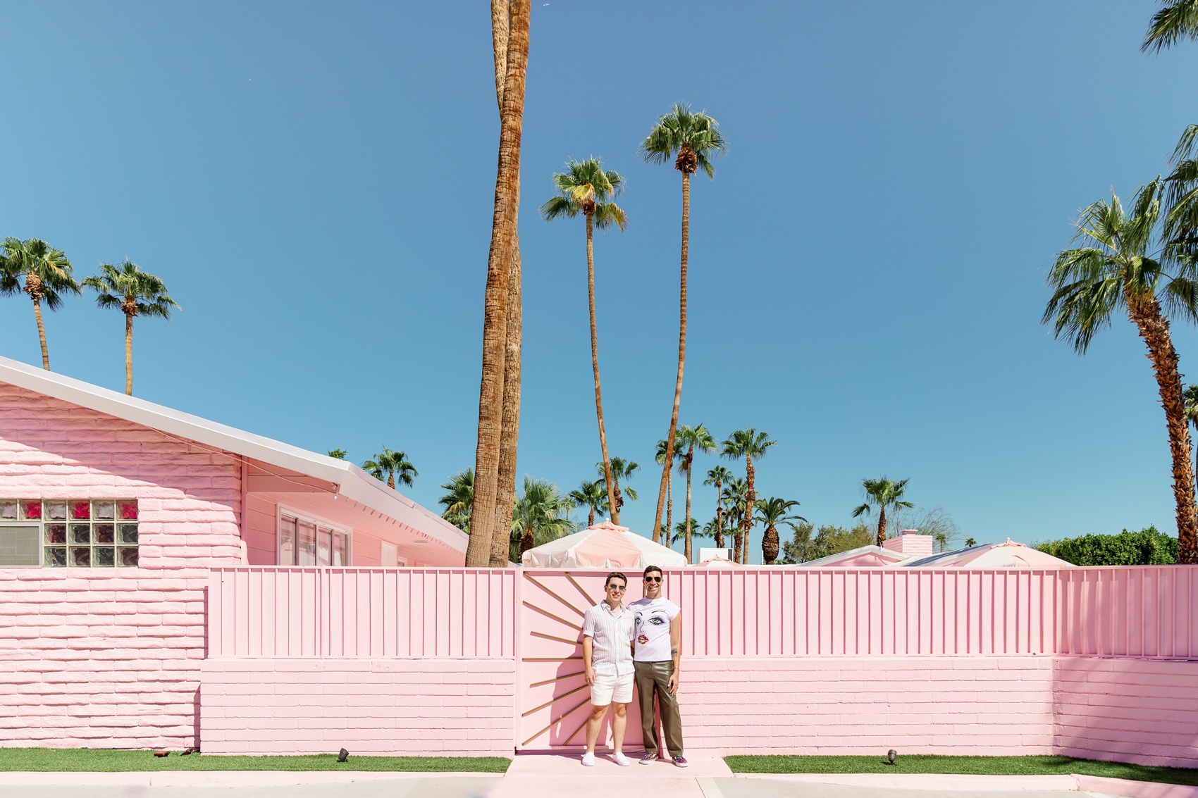 A happy couple stand outside pink walls and gate with big smiles and arms around each other