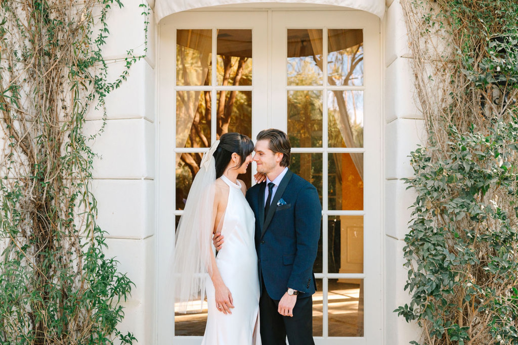 Newlyweds share a snuggle in front of tall glass doors framed in vines at the parker palm springs wedding venue