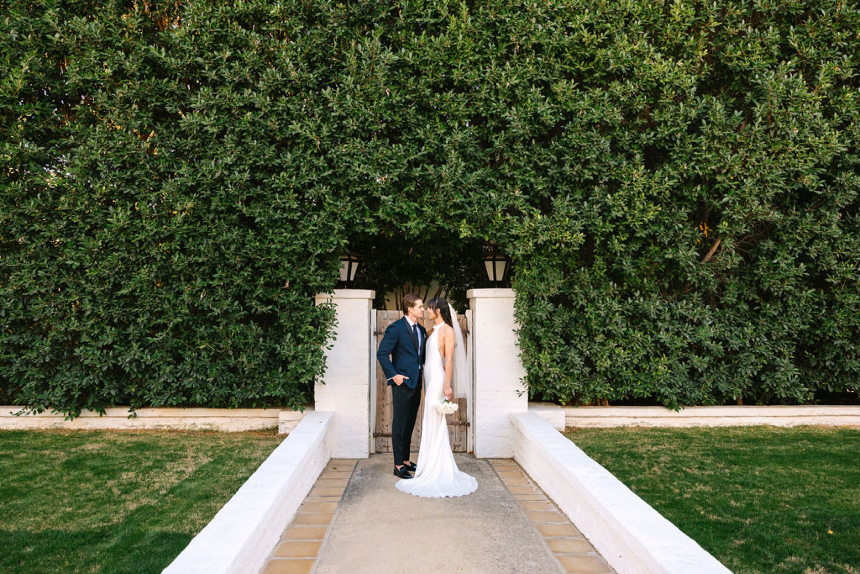 A bride and groom stand smiling at each other in a garden entrance hidden by tall bushes at the parker palm springs wedding venue
