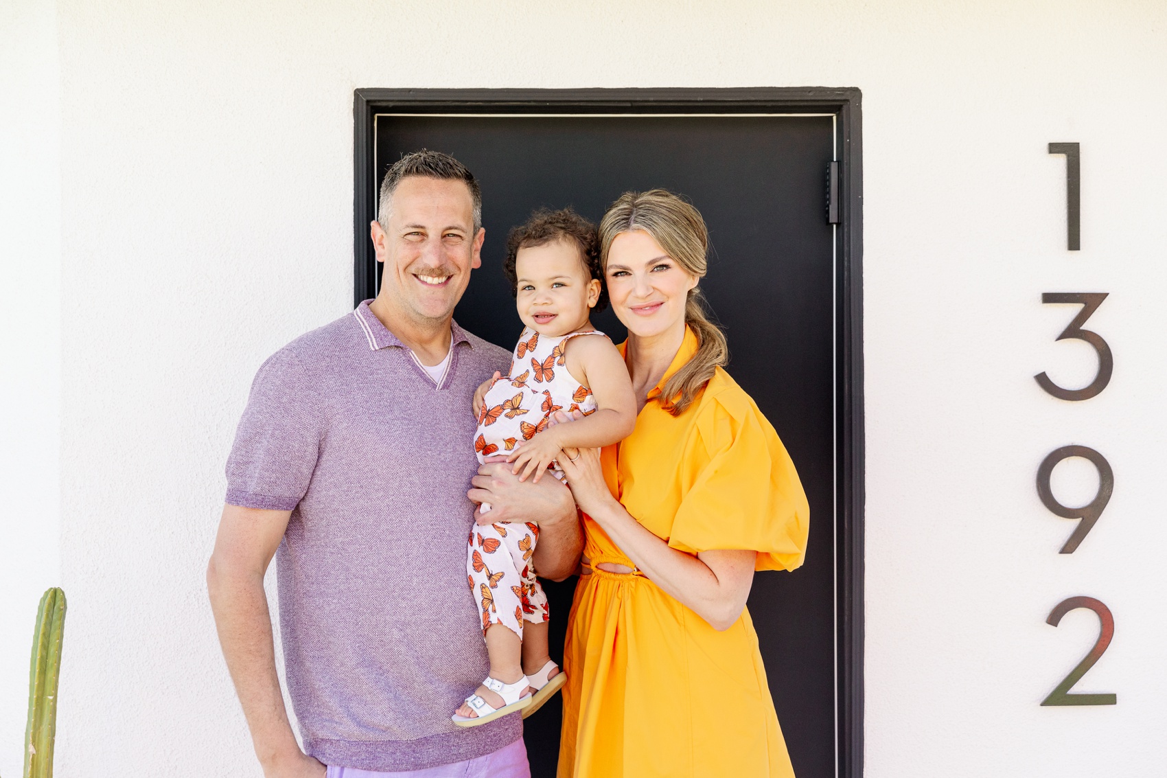 Happy mom and dad stand at their front door holding their toddler daughter between them for their Palm Springs family photographer