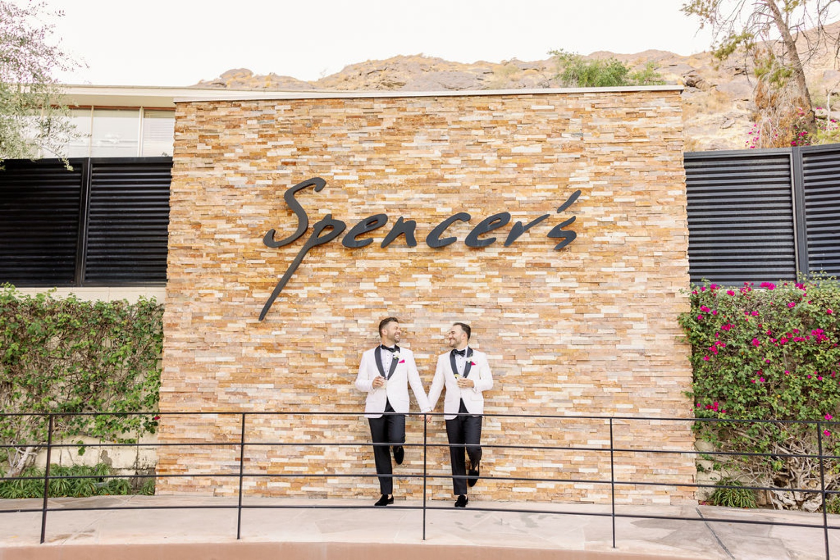 Happy grooms smile at each other while holding hands and leaning against the stone wall of the spencer's restaurant palm springs wedding venue in white tix jackets and black pants