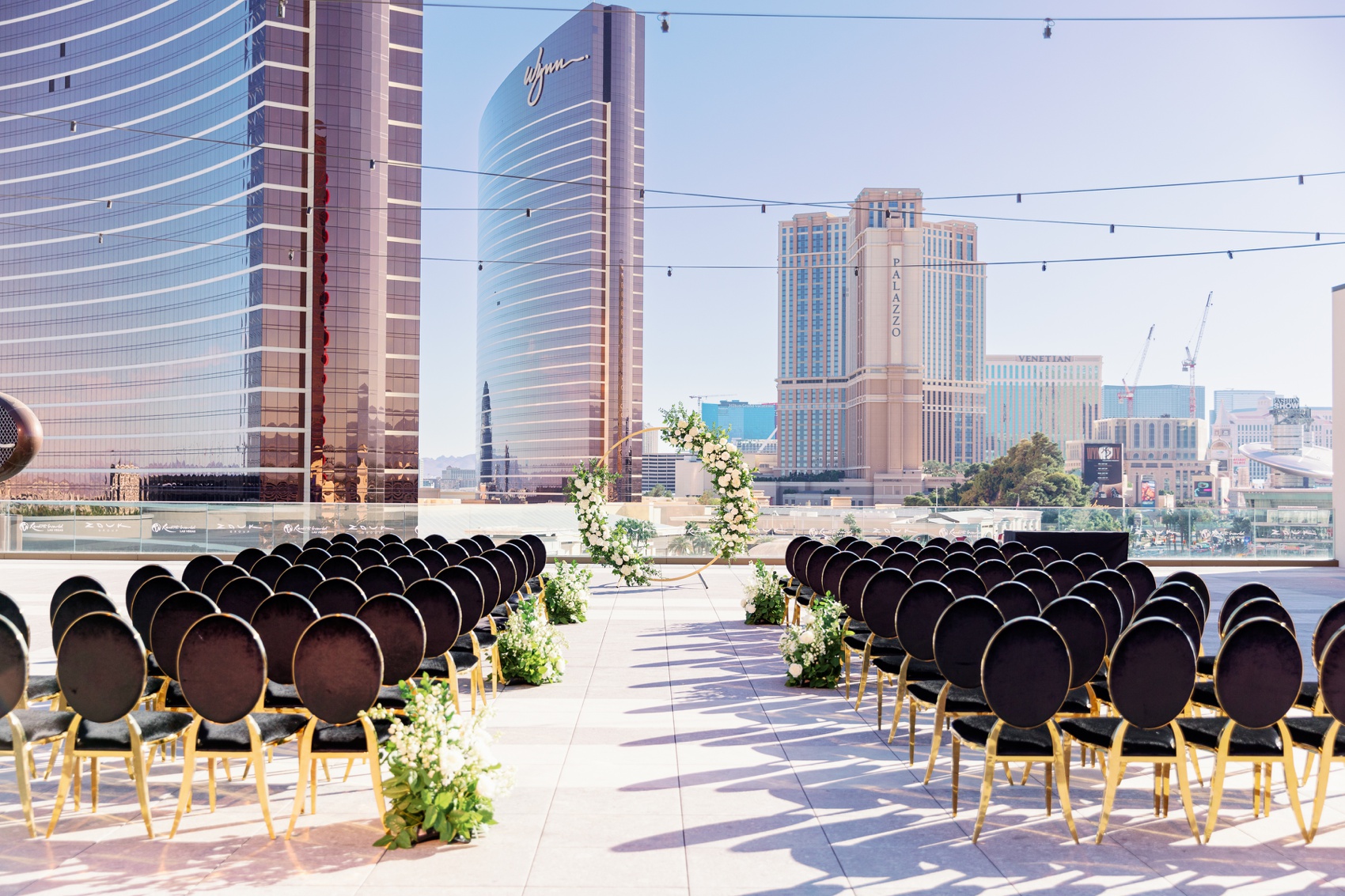 a look down the aisle of a set up wedding reception in las Vegas on a rooftop with black and gold chairs and a white rose floral arch
