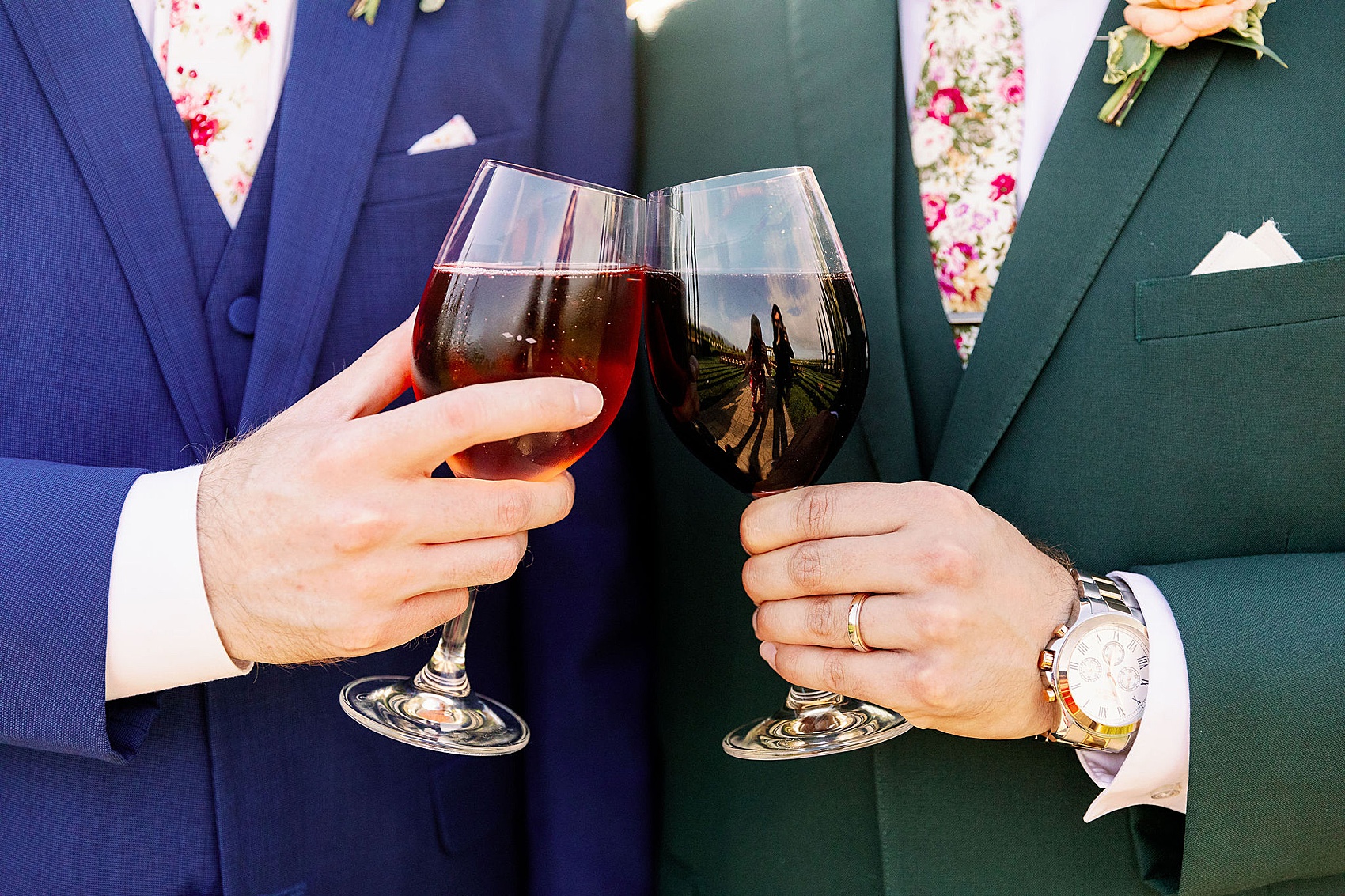 Details of groom toasting drinks in wine glasses against their blue and green suits and matching floral ties