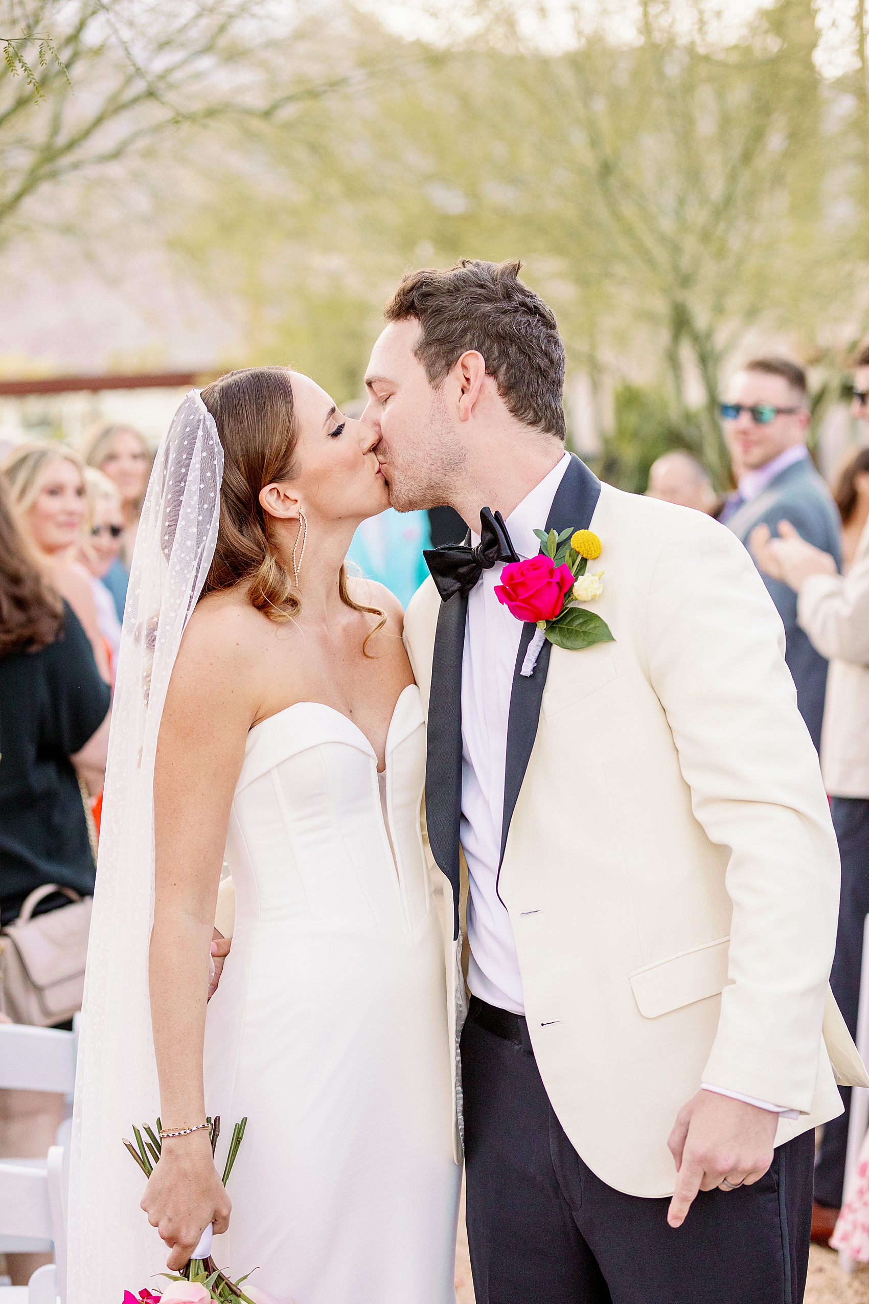 A bride and groom in a white tux jacket kiss in the aisle as guest clap