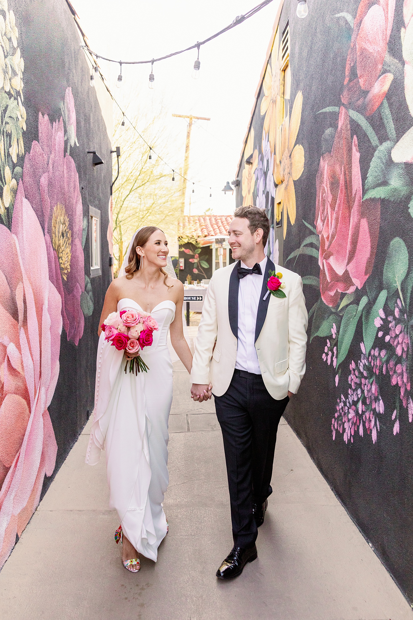 A bride and groom walk holding hands smiling at each other in an alley with murals at Fleur Noire Hotel