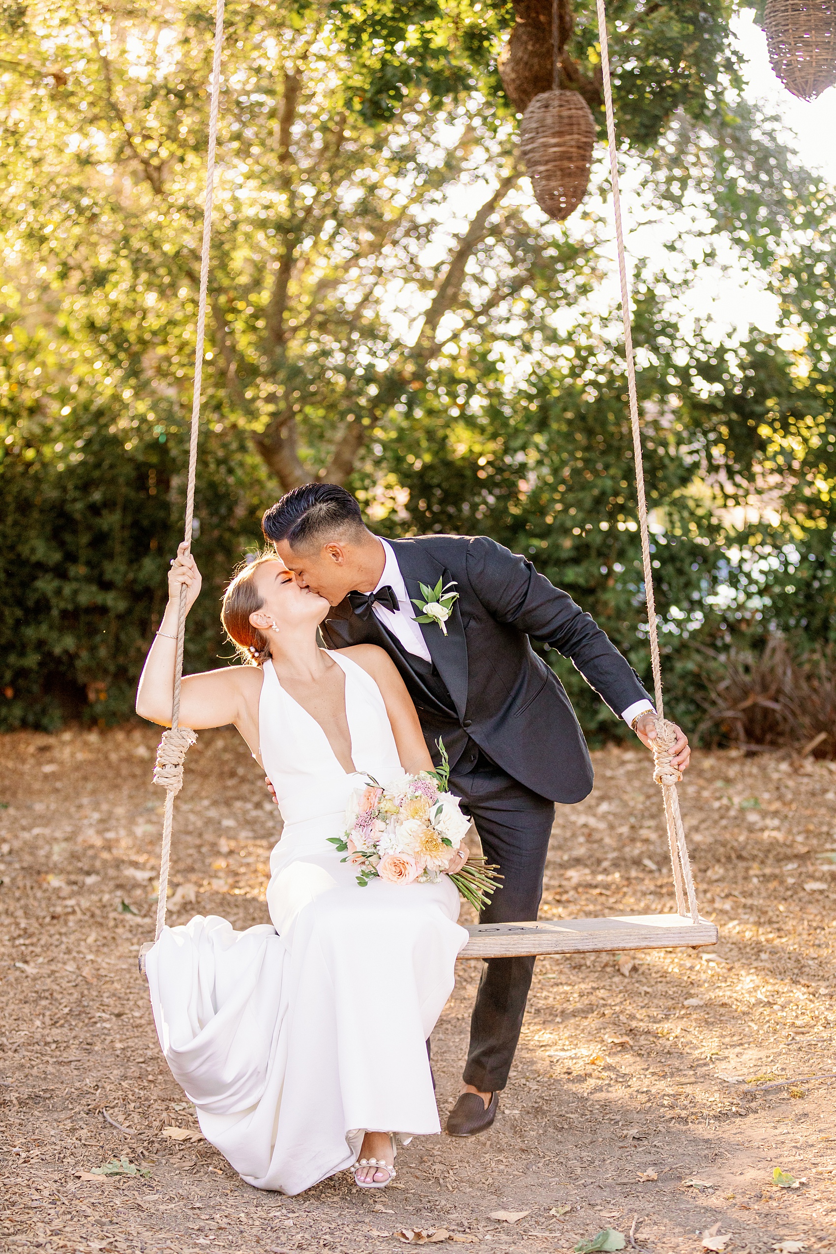 A bride and groom kiss while the bride sits on a rustic swing in a garden at the lodge at sonoma wedding venue