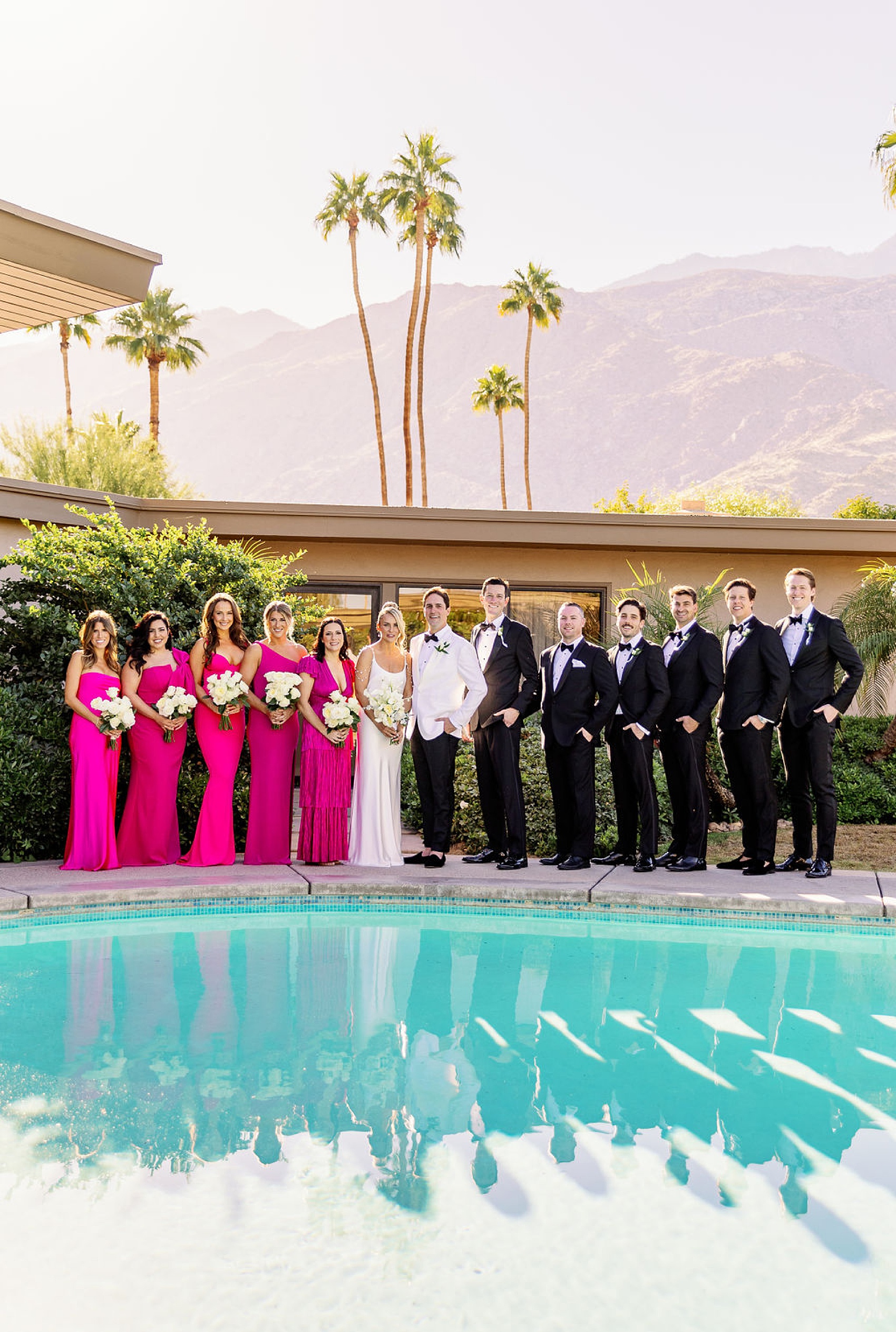 Newlyweds stand with their wedding party in black and pink by the pool at sunset