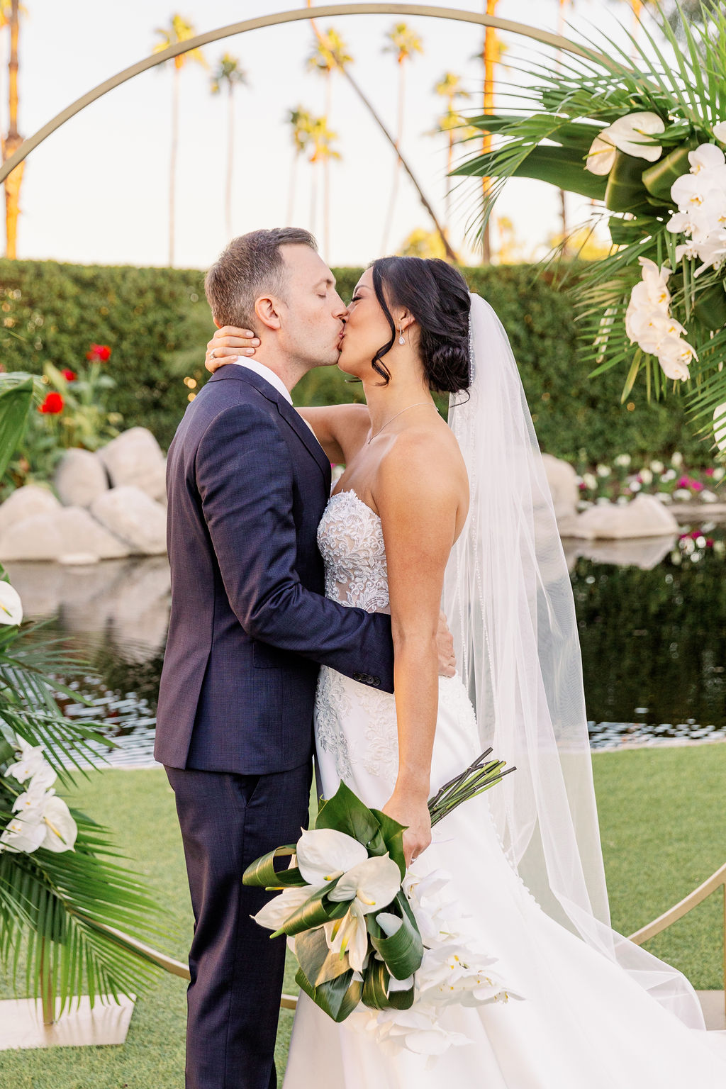 A bride and groom kiss in front of their round gold ceremony arch with white tropical florals at one of the palm springs wedding venues by the water