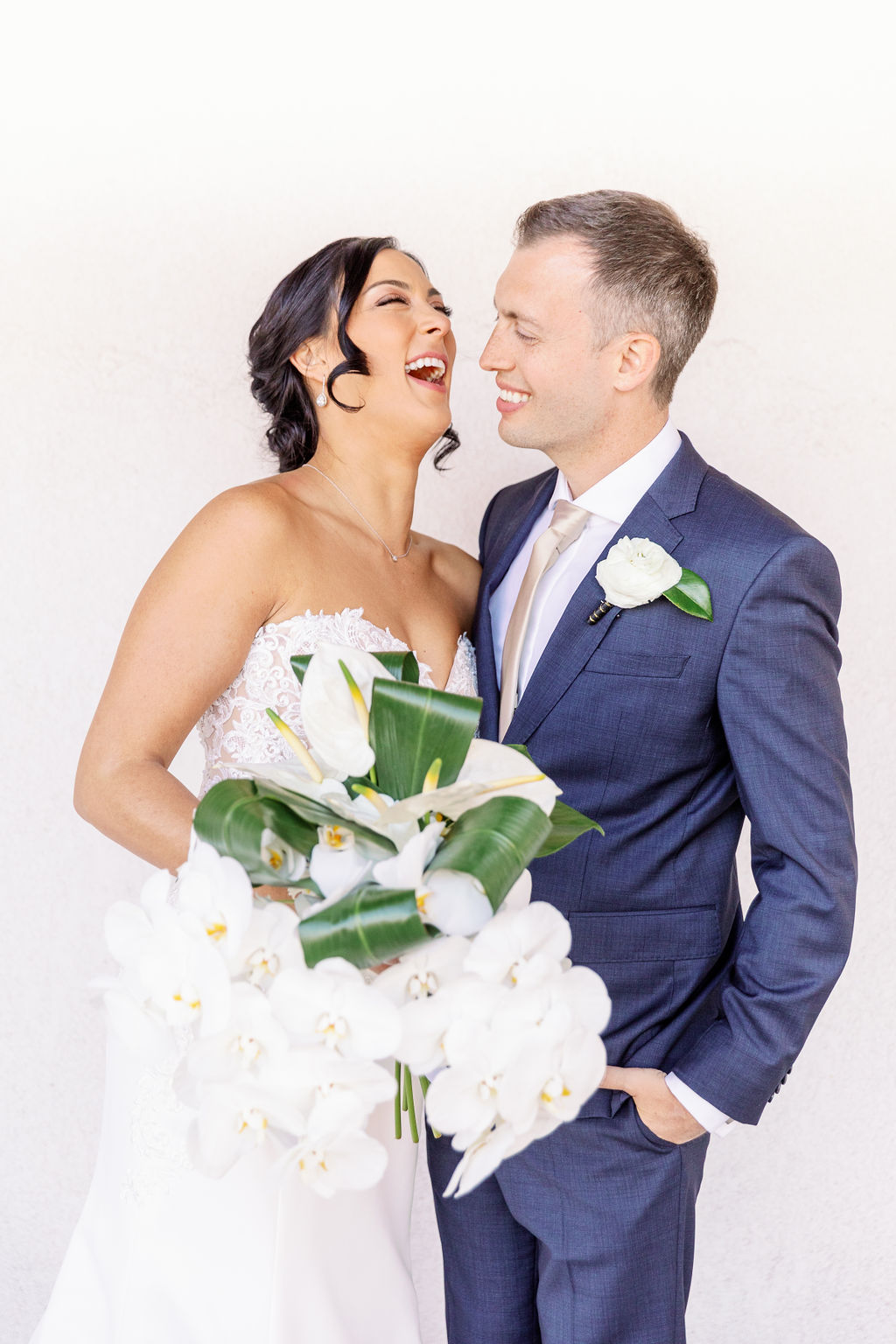 A bride and groom laugh while standing by a white wall holding the large white orchid bouquet at one of the palm springs wedding venues