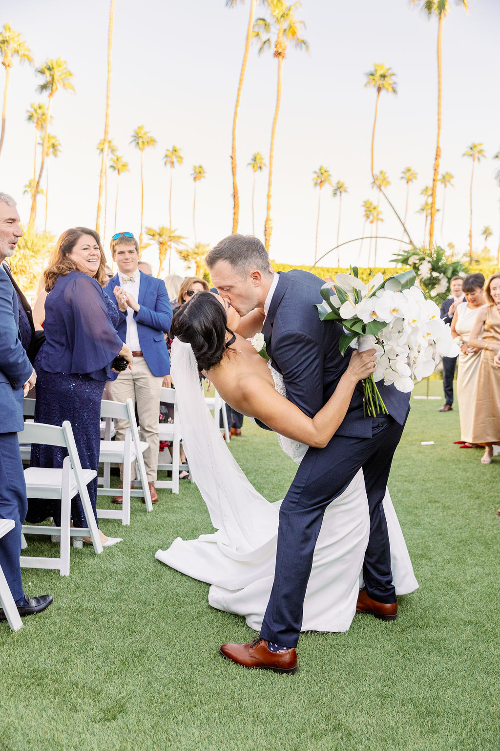 A groom dips his bride in the aisle for a kiss as guests clap at their outdoor ceremony in the lawn of one of the palm springs wedding venues