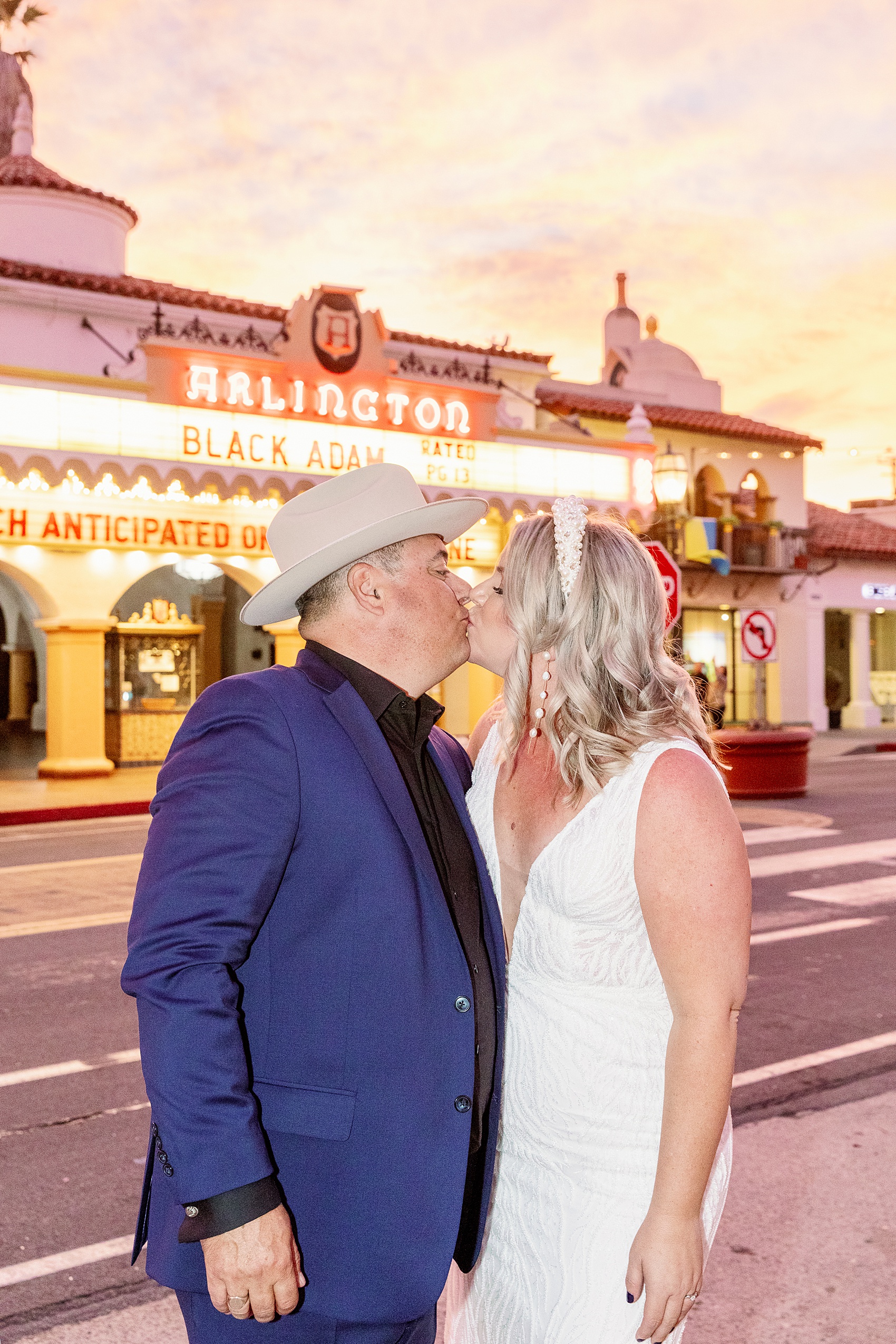 A groom in a blue suit kisses his bride on the sidewalk at sunset in front of a theatre