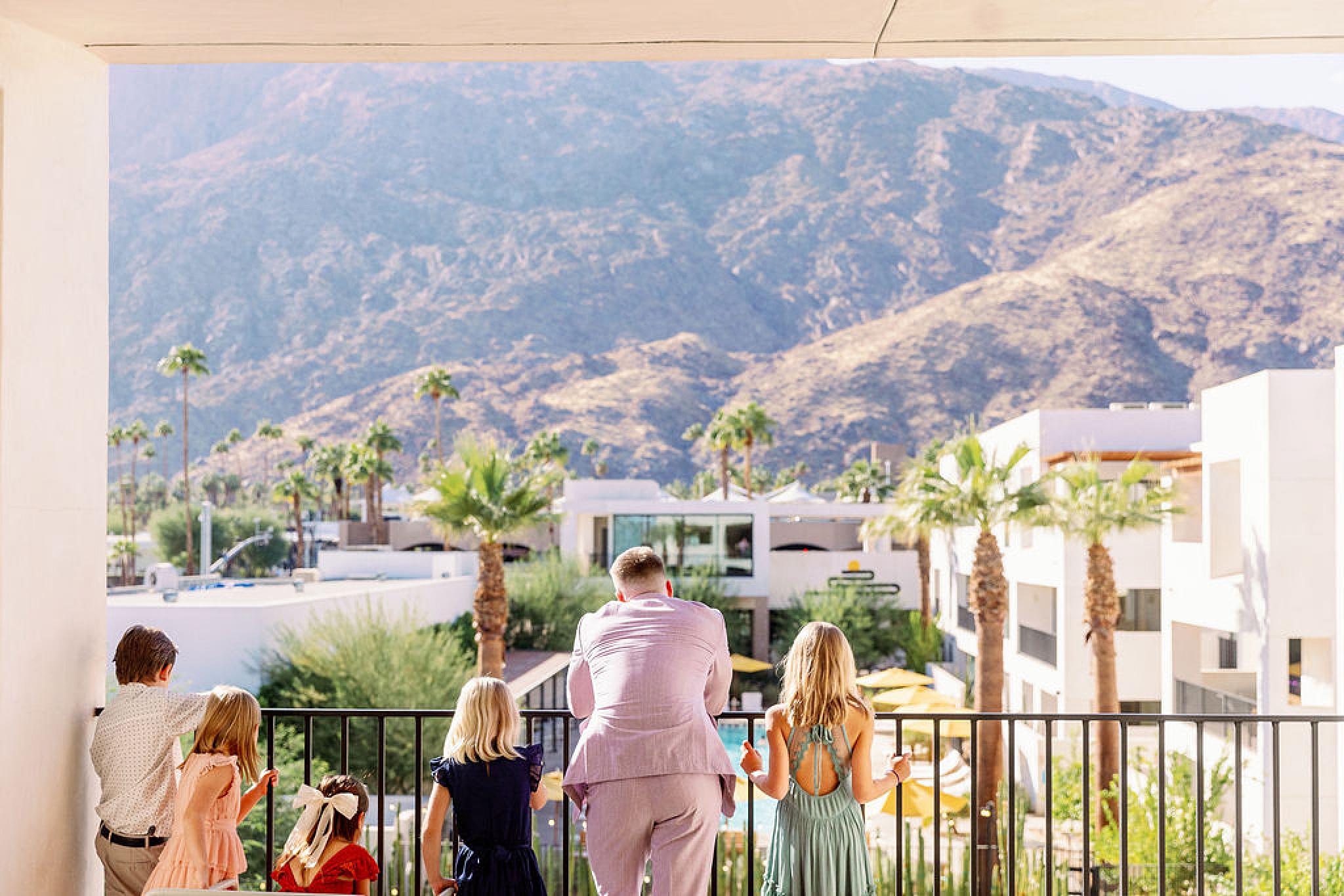 A groom in pink leans against a balcony railing with a group of children overlooking a pool at the drift hotel palm springs wedding venue