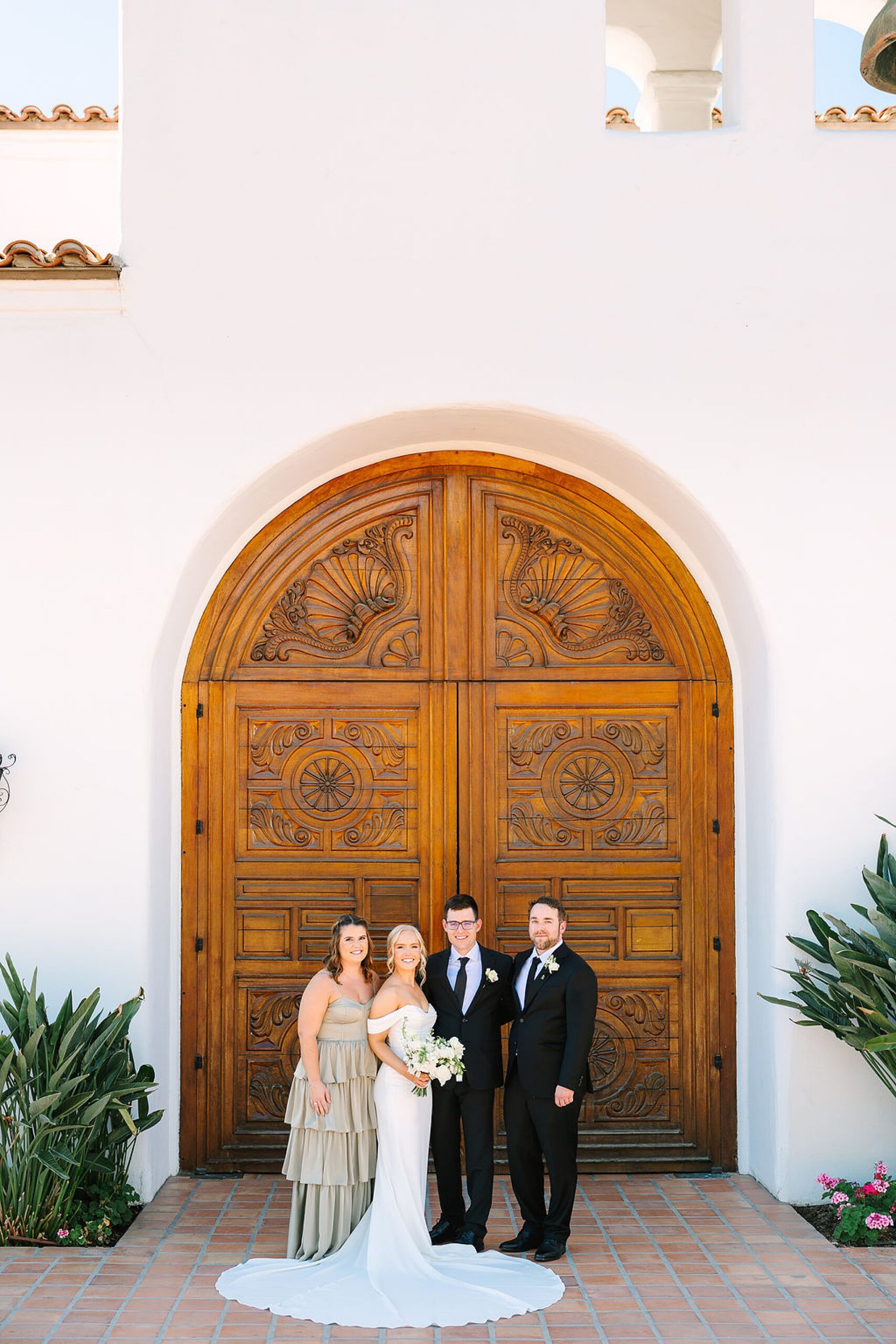 A bride and groom stand with their best man and maid of honor in front of ornate wooden doors