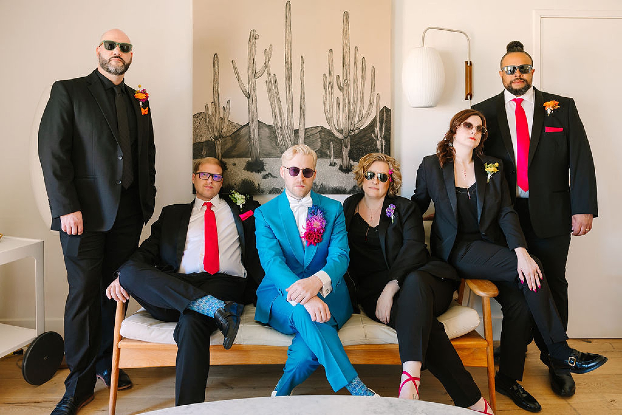 A groom in blue sits in sunglasses on a couch with his wedding party in black with sunglasses as well