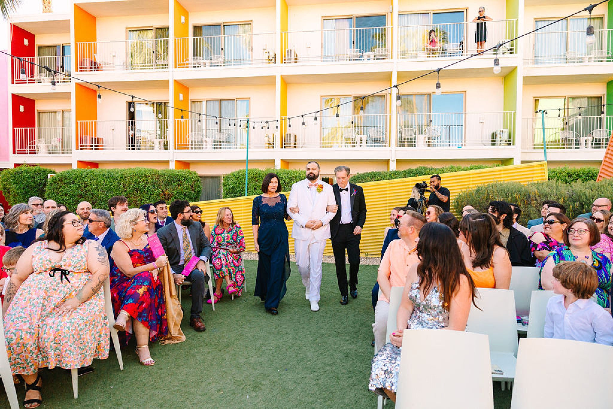 A groom in white is walked down the aisle by mom and dad as guests turn to see